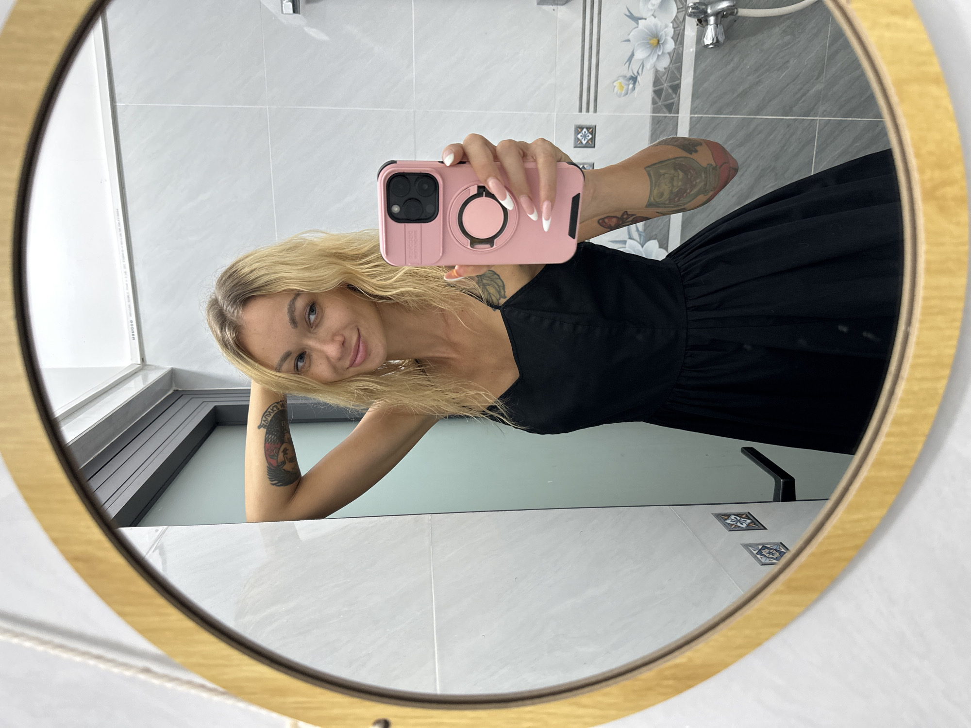 A woman with blonde hair and fair skin takes a mirror selfie in what appears to be a bathroom in Mũi Né, Vietnam. She is wearing a black dress and holding a pink iPhone with a ring stand in her right hand. Her fingernails are painted white with French tips. She has tattoos on both arms. The mirror is oval-shaped with a golden frame. In the reflection, the bathroom has light-colored tiled walls and a shower or tub area. There is a faucet visible. The overall lighting suggests daytime. The woman's expression is a slight smile.