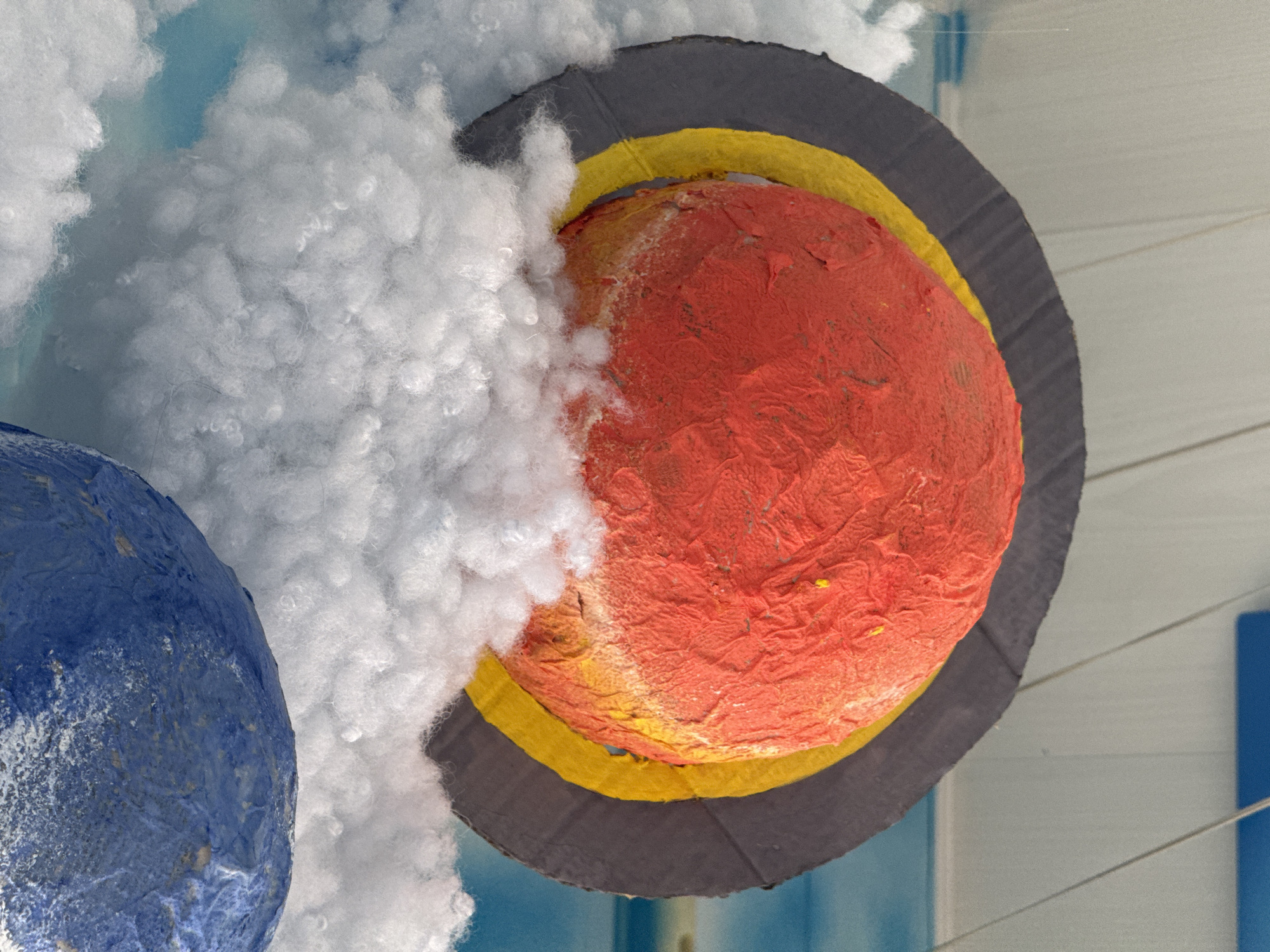 The image shows two constructed planets hanging from a ceiling, likely as part of a decoration or display. One planet is red with a yellow ring around it, surrounded by a larger gray ring. The other planet is blue. Both are partially obscured by fluffy white material that resembles clouds. The background is a light blue with vertical white and blue bars. Based on metadata, the location is Kladovitsy, Russia. No people are visible. The focus is on the crafted planetary models.