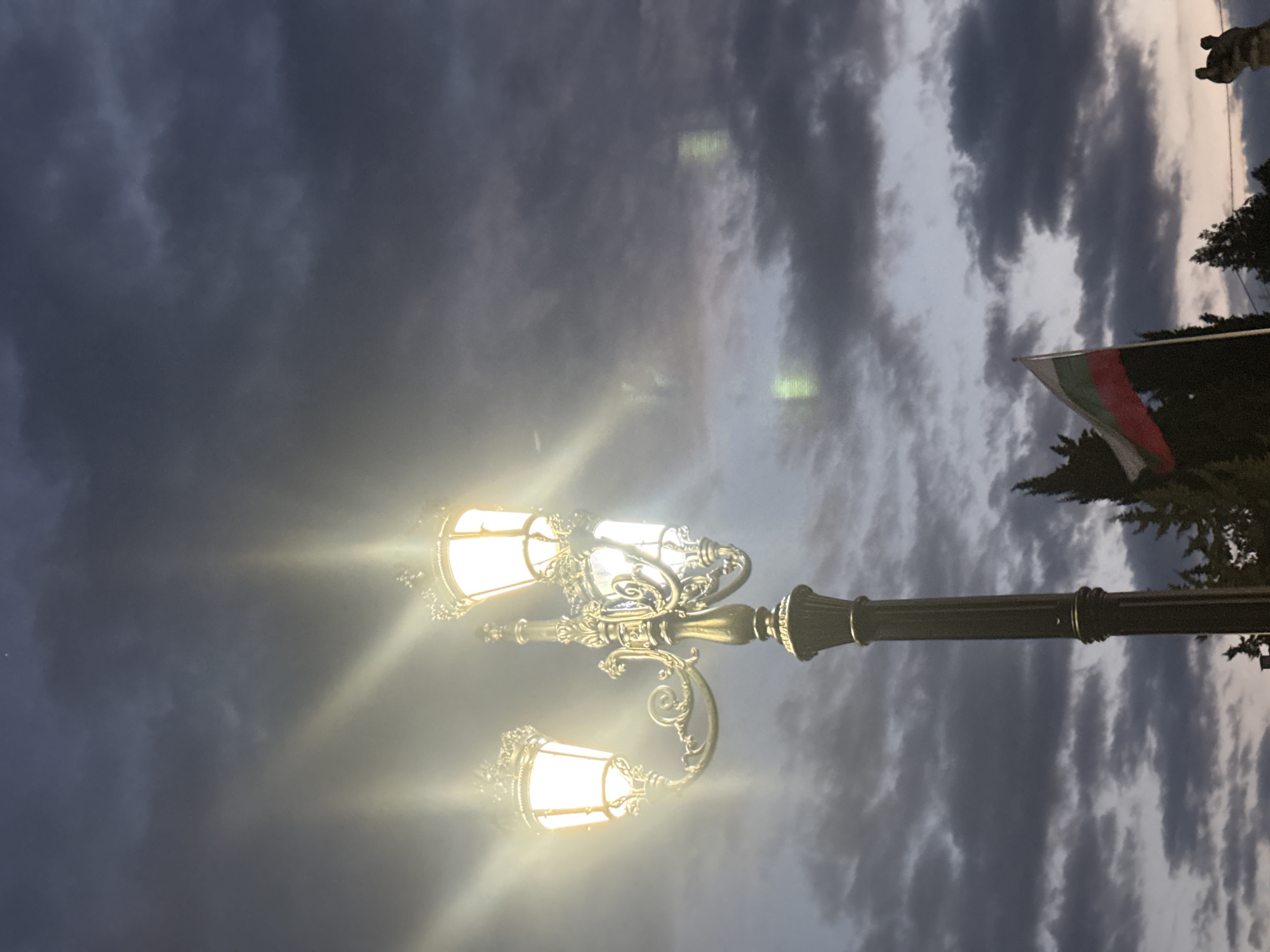 The image, captured in the city of Strelcha, Bulgaria, depicts an ornate street lamp with three illuminated globes, casting a bright glow against a cloudy twilight sky. The lamp is a metallic structure with intricate detailing. To the right of the lamp, a Bulgarian flag is visible, fluttering on a pole. The background is a dark, overcast sky with thick clouds, suggesting an evening or pre-storm atmosphere. Silhouetted trees are visible at the bottom of the frame, partially obscuring the base of the lamp post.