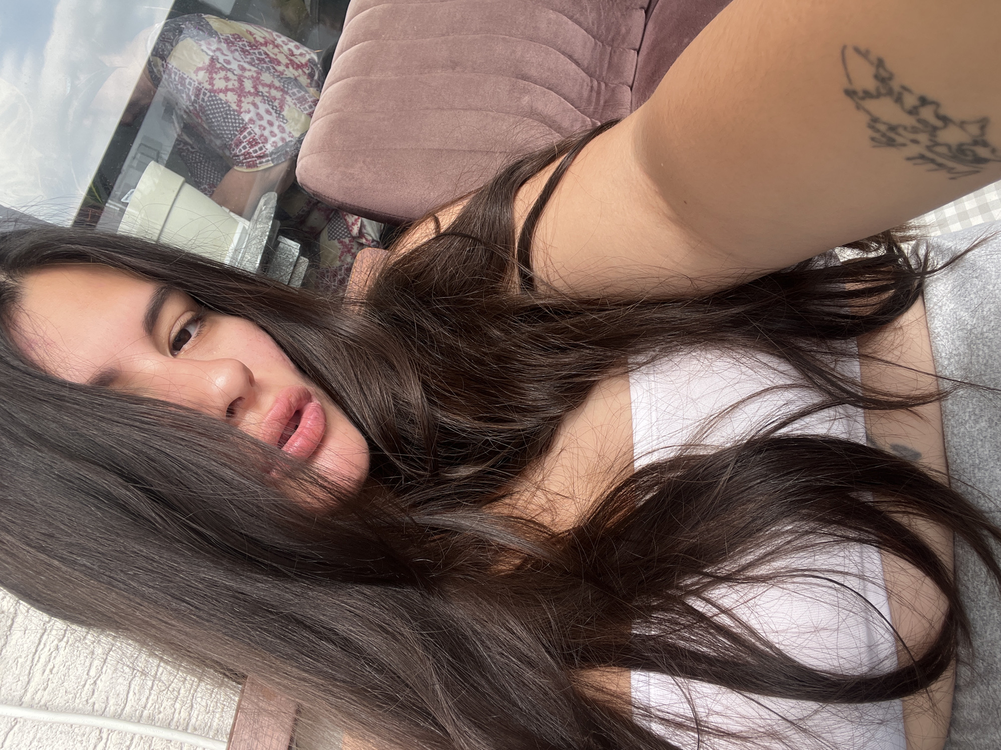 A woman is lying on her side, partially visible, wearing a white top. She has long, dark hair that partially covers her face. Her left arm is raised, revealing a tattoo. The background features a blurred reflection in a window, showing elements of an interior with seating. Location: unspecified area within Kiryat Bialik, Israel.