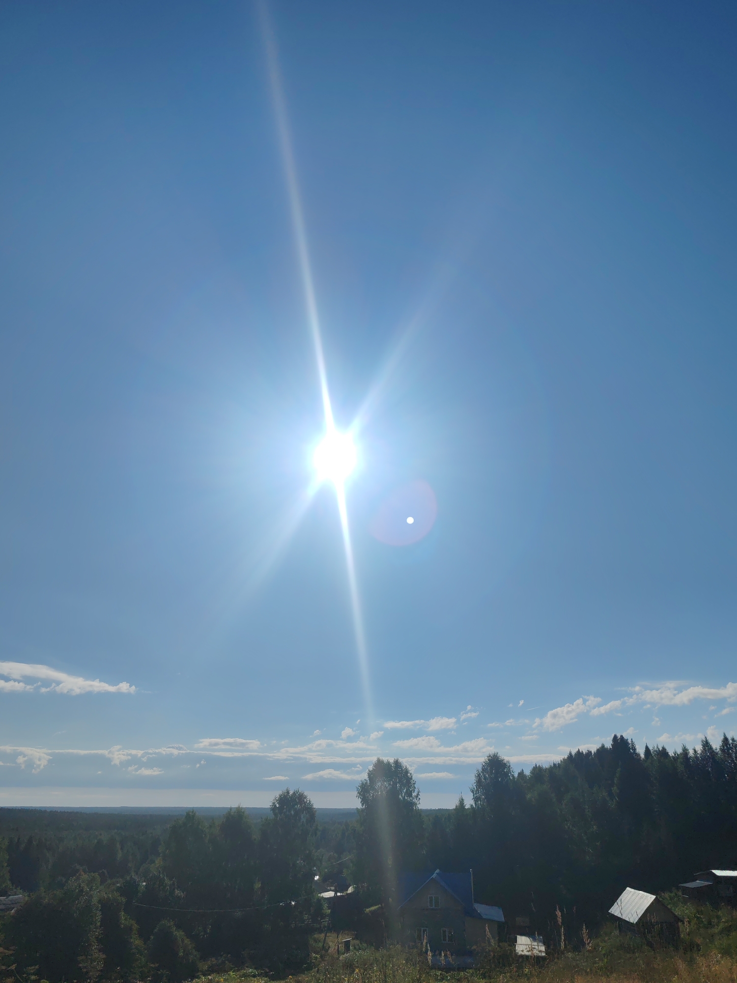 The image depicts a bright, sunny day in a rural landscape, likely in or near the settlement of Chukhlem, Russia. The sky is a clear, vibrant blue, dominated by the intense glare of the sun, which is positioned high in the frame, creating prominent sunburst effects and lens flares. A few wispy white clouds are scattered across the horizon. Below the sky, a dense, dark green forest stretches across the mid-ground, with the tops of the trees silhouetted against the lighter sky. In the foreground and scattered amongst the trees, there are several small houses with visible roofs, suggesting a residential area or small village. The vegetation in the foreground is also green, with some dry grasses visible. The overall impression is of a natural, undeveloped landscape under a bright, midday sun.
