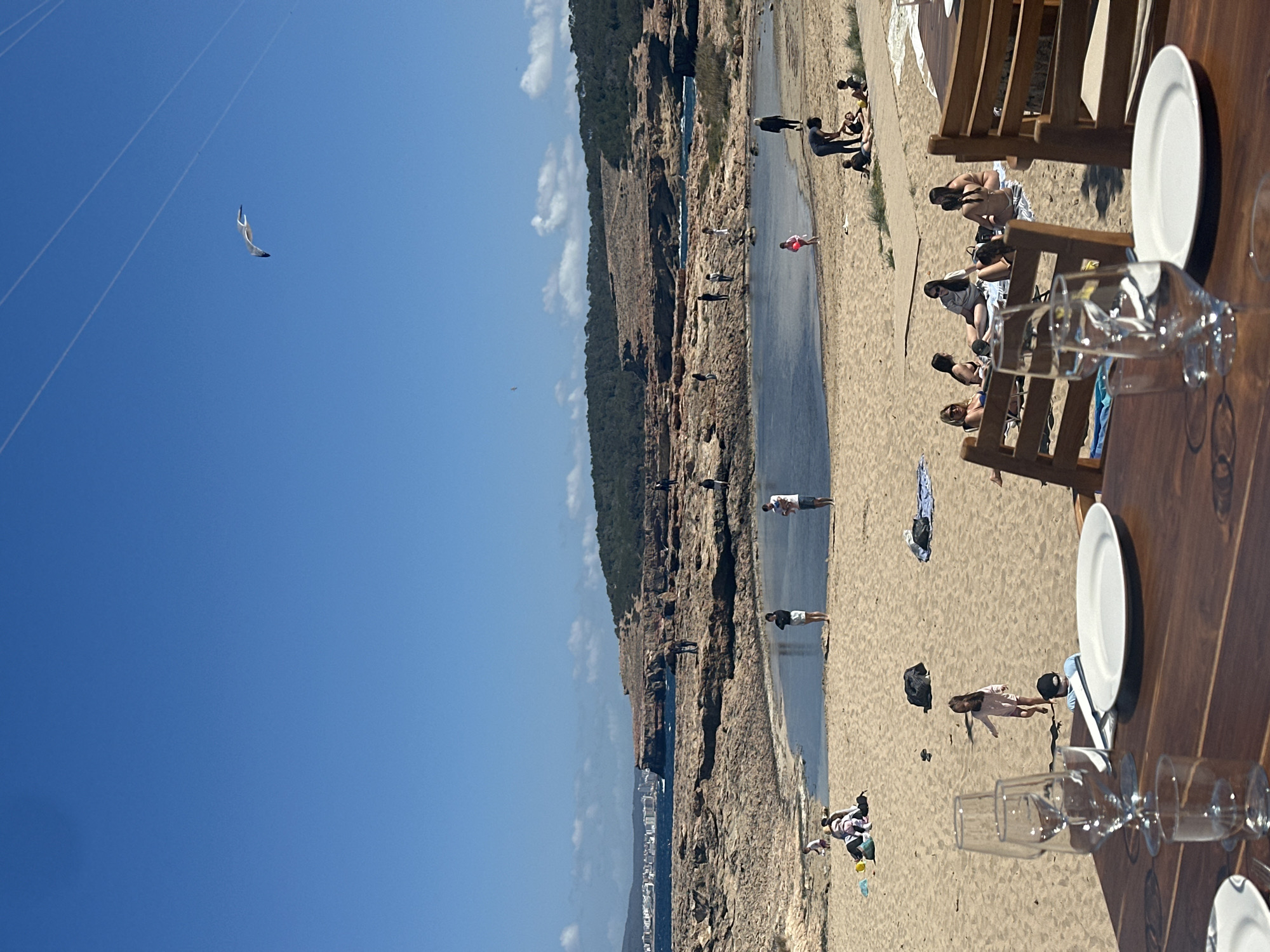 The scene depicts a beach in Sant Josep de sa Talaia, Spain, likely along the western coast given the perspective. The image is taken from an elevated viewpoint, possibly from a restaurant or terrace overlooking the beach.
In the foreground, a wooden table is set with plates and glasses, indicating a dining area. Beachgoers are present on the sandy beach below. Some people are sunbathing or relaxing on chairs, while others are walking along the waterline. The presence of umbrellas and beach towels suggests leisurely activities. A rocky coastline borders the beach, and lush greenery can be observed on top of the cliffs. In the distance, the coastline extends into the horizon.
The sky is clear and blue with some scattered clouds. High above, a paraglider or kite surfer is visible. The presence of support wires suggests the paraglider is attached to a winch or similar device, likely part of a water-based activity.