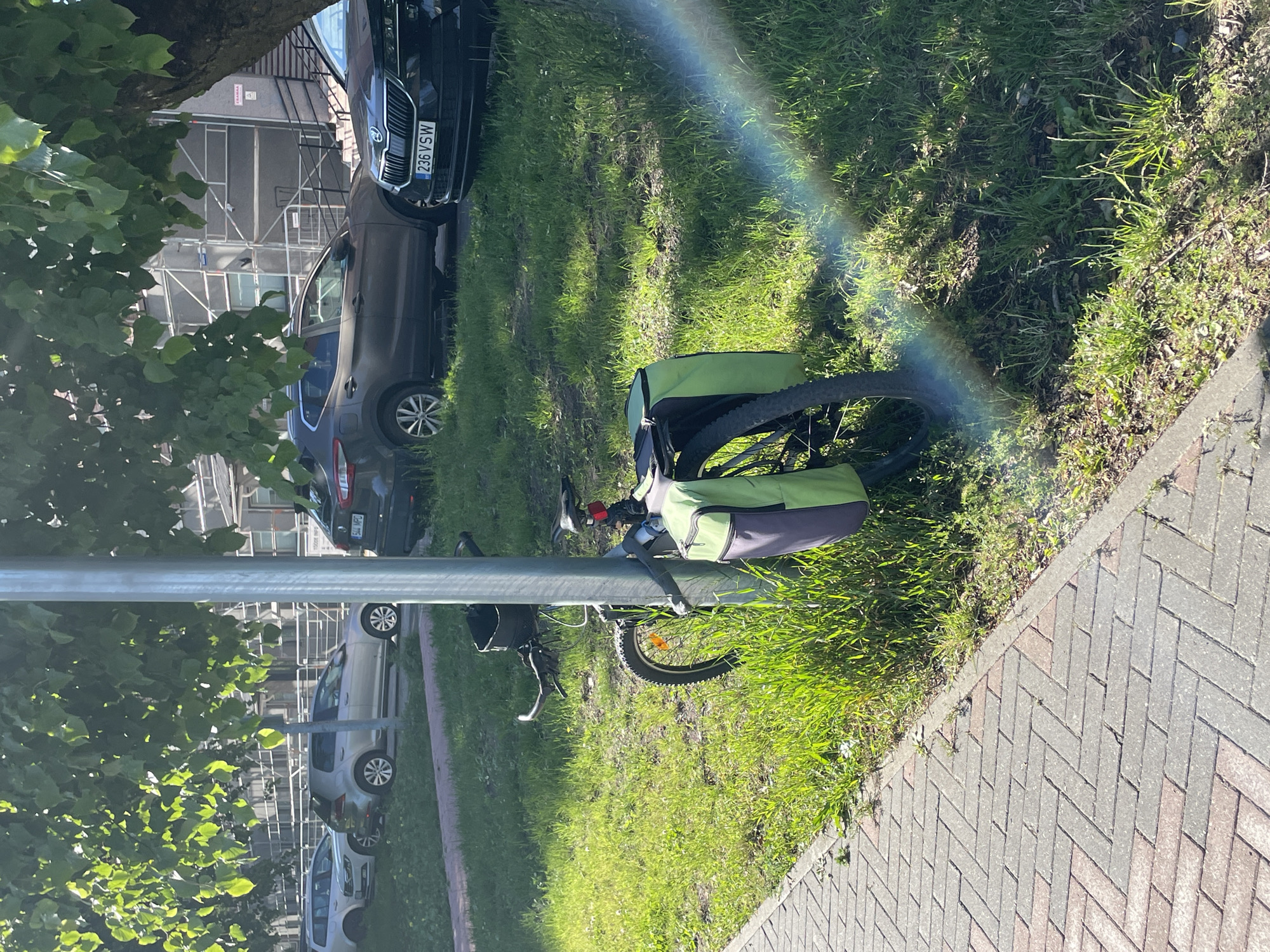 The image shows a bicycle parked next to a street in Tallinn, Estonia. The bicycle has two green panniers attached to its rear rack. A rainbow effect is visible in the upper right portion of the image, suggesting it might be sunny and raining simultaneously. Two cars are parked behind the bicycle on the grassy verge. One of the cars has a license plate that reads "236 VSW". In the background, there are buildings under construction with scaffolding. The image appears to have been taken during the daytime, with bright sunlight illuminating the scene. The overall mood is peaceful, with the bicycle appearing to be temporarily stored while its owner is engaged in other activities.