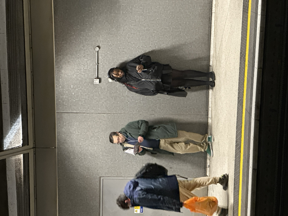 The image displays an indoor train platform, characteristic of the London Underground or Overground network in the city of London, United Kingdom. The platform features walls clad in small, light grey square tiles, and a concrete floor. A distinct yellow safety line, bordered by a black line, runs along the platform edge, above the dark train track area. An overhead metal structure with a translucent or textured panel is visible at the top.
Three individuals are present. On the far left, a person in a dark blue hooded jacket and light-colored trousers is in motion, walking from left to right. This individual carries a black backpack on their left shoulder and an orange plastic shopping bag in their right hand. In the center, a male subject stands stationary, wearing a dark green coat over a blue garment, light brown trousers, and glasses. He is engrossed in reading an open book or magazine held in both hands at chest level, and is wearing white wired earphones. On the right, a female subject stands stationary, leaning against the tiled wall. She wears a black bomber jacket over a dark skirt, black tights, and flat black shoes. Her hair is styled in braids, with some visible reddish strands. She is looking down at a dark smartphone held in both hands and wears black over-ear headphones. A black shoulder bag hangs from her left shoulder. There are no direct interactions observed between the individuals.