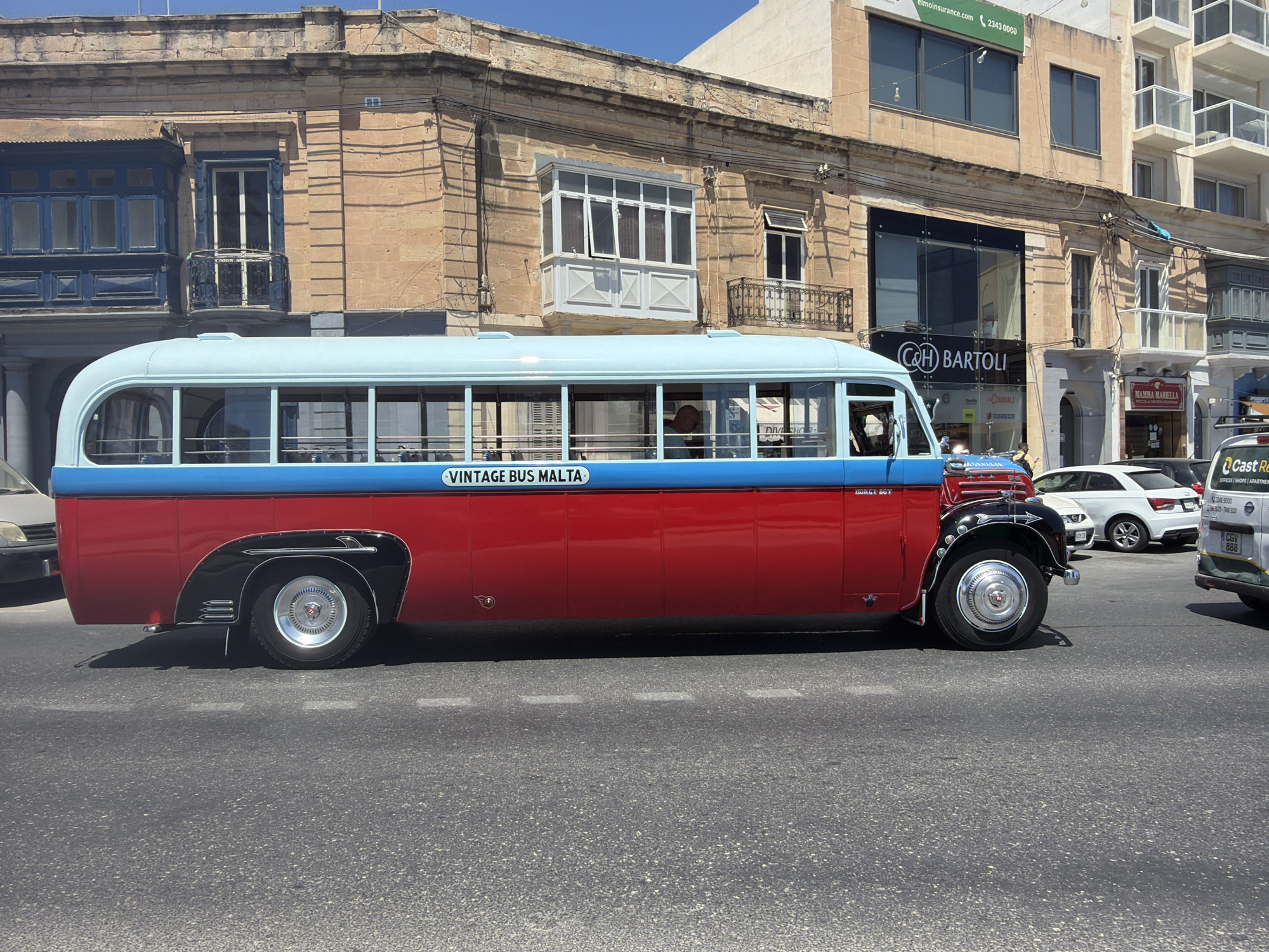 A vintage red and blue bus with "VINTAGE BUS MALTA" written on its side is parked on a street in Gżira, Malta. The bus has large windows and chrome accents. In the background, there are several buildings with balconies and shops. One of the shops has a sign that reads "C&H BARTOLI". Other vehicles, including white cars and a white van, are also visible on the street. The sky is clear and blue.