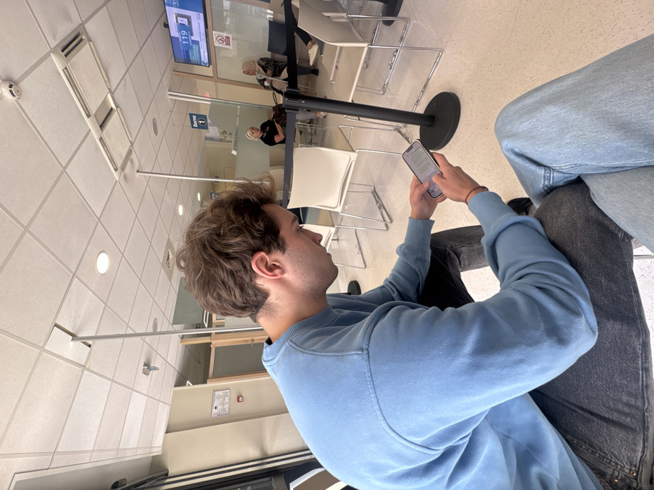 The image depicts an indoor waiting area within a public service office in Saint-Priest, France. In the foreground, a male individual, approximately 20-30 years old with light brown, wavy hair, is seated. He wears a light blue crew-neck sweatshirt and dark grey jeans, and is focused on a light-colored smartphone held with both hands, interacting with the screen. A thin red string bracelet is on his right wrist. The leg of another person, wearing light blue jeans, is partially visible in the bottom right.
The waiting area features a grid-patterned suspended ceiling equipped with recessed circular lighting fixtures, rectangular air vents, and a smoke detector. The floor is a light-colored, speckled tile or linoleum. Transparent glass partitions with vertical metal supports delineate sections of the space. Black stanchions with retractable belts are present for queue control.
In the mid-ground, two women are seated in white, contemporary-style chairs with metal frames. One older female, with blonde hair and a black top, is positioned center-left, with a dark bag on an adjacent chair. Another female, wearing a headscarf and a patterned top, is seated further right.
On the far right wall, a digital queuing system display shows "119" as the current number being served, with an associated counter "25" indicated below. A red circular 'No Smoking' emblem is affixed to the wall beneath the display. A sign marked "Borne 1" (Kiosk 1) is mounted above a glass partition. A red-bordered informational sign is affixed to the left wall. The scene suggests a typical public administrative or service waiting room environment.