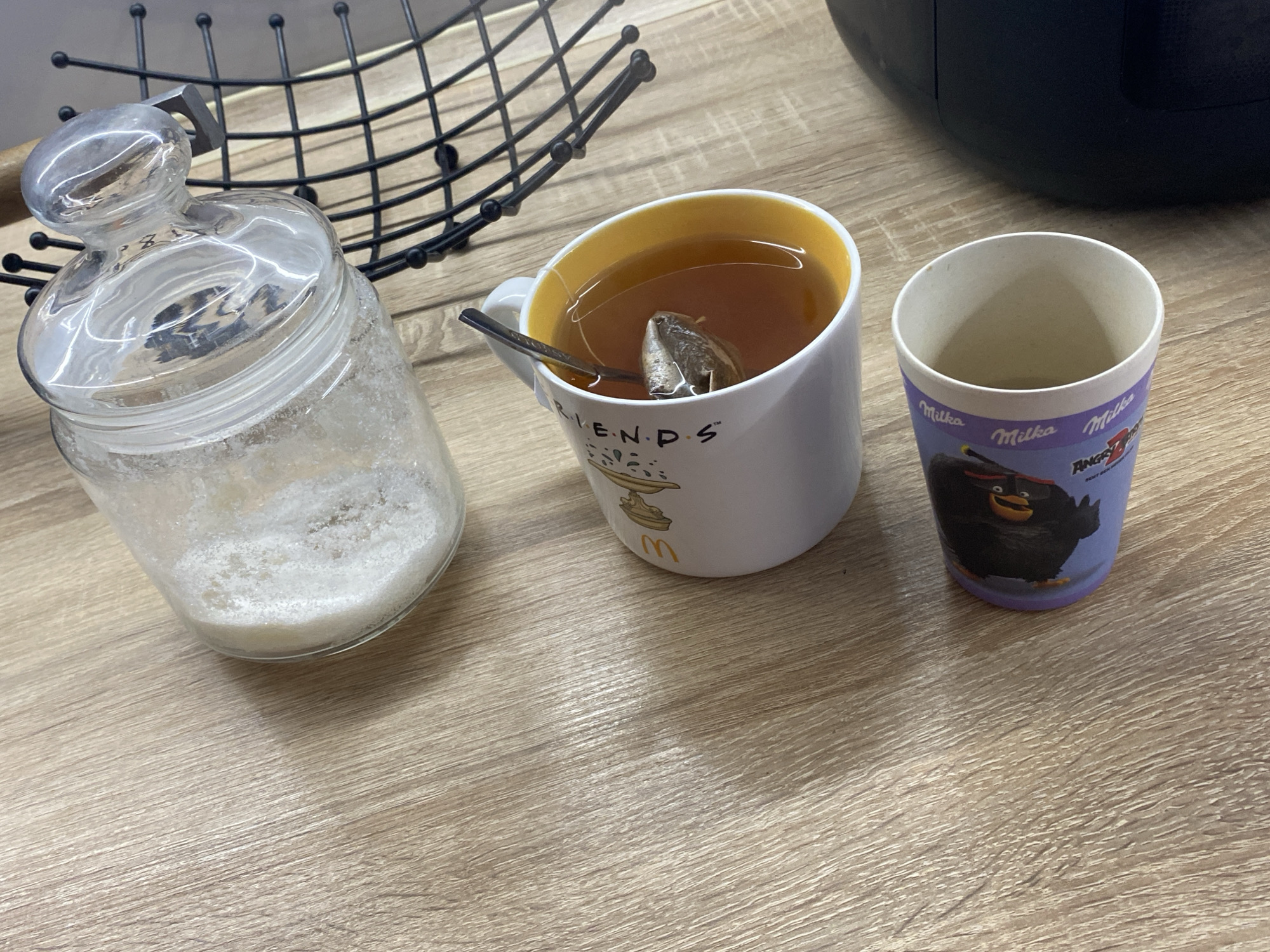 The image depicts a close-up, high-angle shot of a wooden table with three items: a glass jar containing white granular material, a white mug with a yellow interior filled with tea and a tea bag, and a small purple cup with a cartoon character on it. The glass jar has a clear glass lid and appears to be a sugar or salt shaker. The mug has text that appears to read "FRIENDS" and a McDonald's logo. The purple cup has "Milka" branding and an Angry Birds character. In the background, a portion of a black metal rack is visible. The overall scene is brightly lit, suggesting an indoor setting. There are no people visible in the image. This scene is located in Kryvyi Rih, Ukraine.