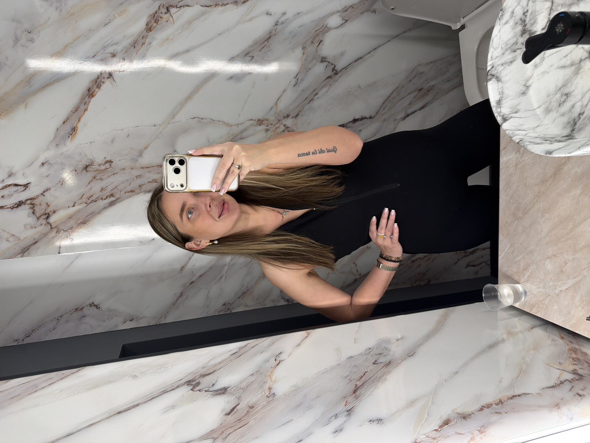 A woman is taking a selfie in a bathroom. She is wearing a black outfit and has blond hair. The bathroom has white marble walls and a countertop with a sink. A plastic cup filled with water sits on the countertop. The location is not discernible from the image, so the assertion that it is in Moscow, Russia, cannot be validated.