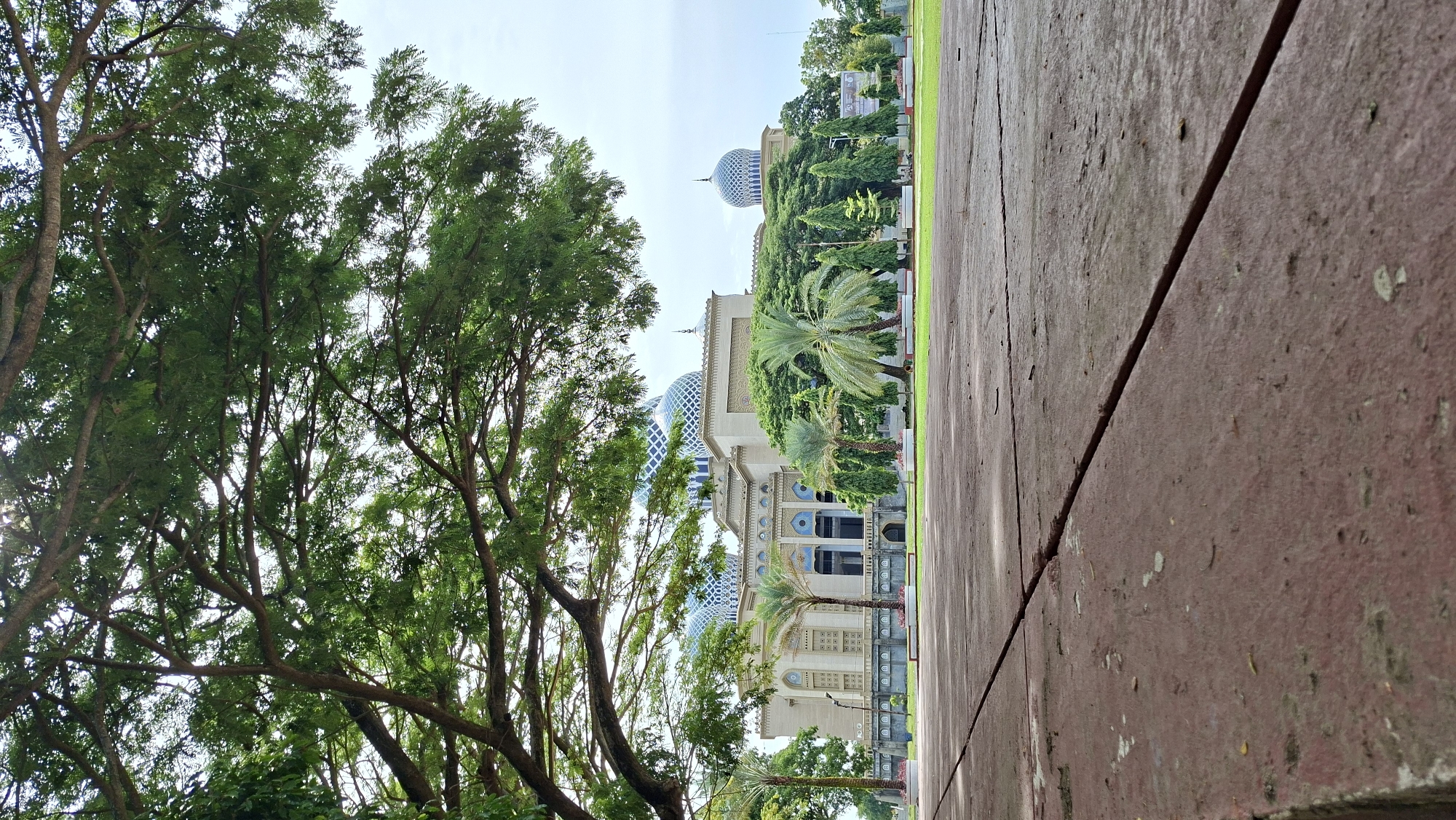 The image shows the Baiturrahman Grand Mosque in Lhokseumawe, Indonesia, viewed from a low angle. The mosque features a large central dome and several smaller domes, all decorated with intricate blue and white patterns. The building is light-colored with arched windows and decorative elements. In front of the mosque is a manicured lawn with several palm trees and other greenery. The foreground is dominated by a reddish-brown paved area with visible cracks. Large trees with lush green leaves partially obscure the view of the sky and the upper parts of the mosque, framing the scene from the left. The sky is overcast. There are no people visible in the image.