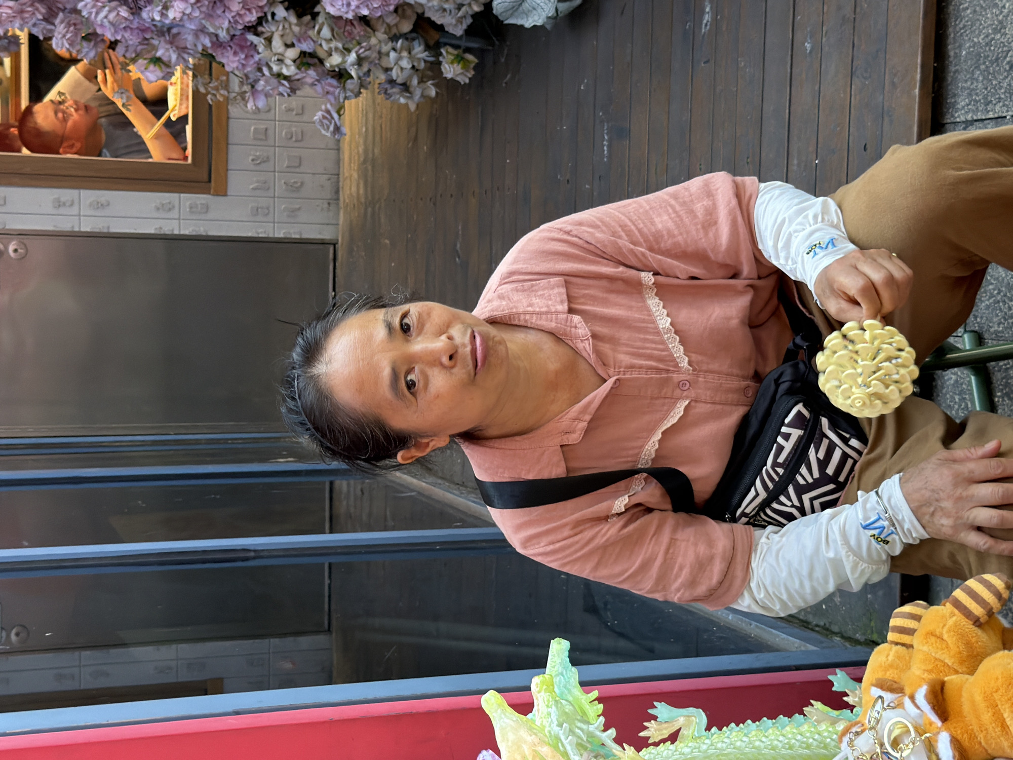 A woman sits outdoors on a wooden deck, holding a yellow, flower-shaped wafer in her right hand. She wears a pink, button-down shirt with white lace trim at the collar and cuffs, layered over a white long-sleeved shirt. A black fanny pack with a zigzag pattern is worn across her chest. She has dark hair pulled back and is looking towards the camera with a neutral expression. In the background, a man is visible through a window, looking at his phone and holding chopsticks. To the left of the woman, part of a green, dragon-like sculpture is visible. The overall environment appears to be a street or market area in Yuzhong District, China, with a tiled wall and wooden floor.