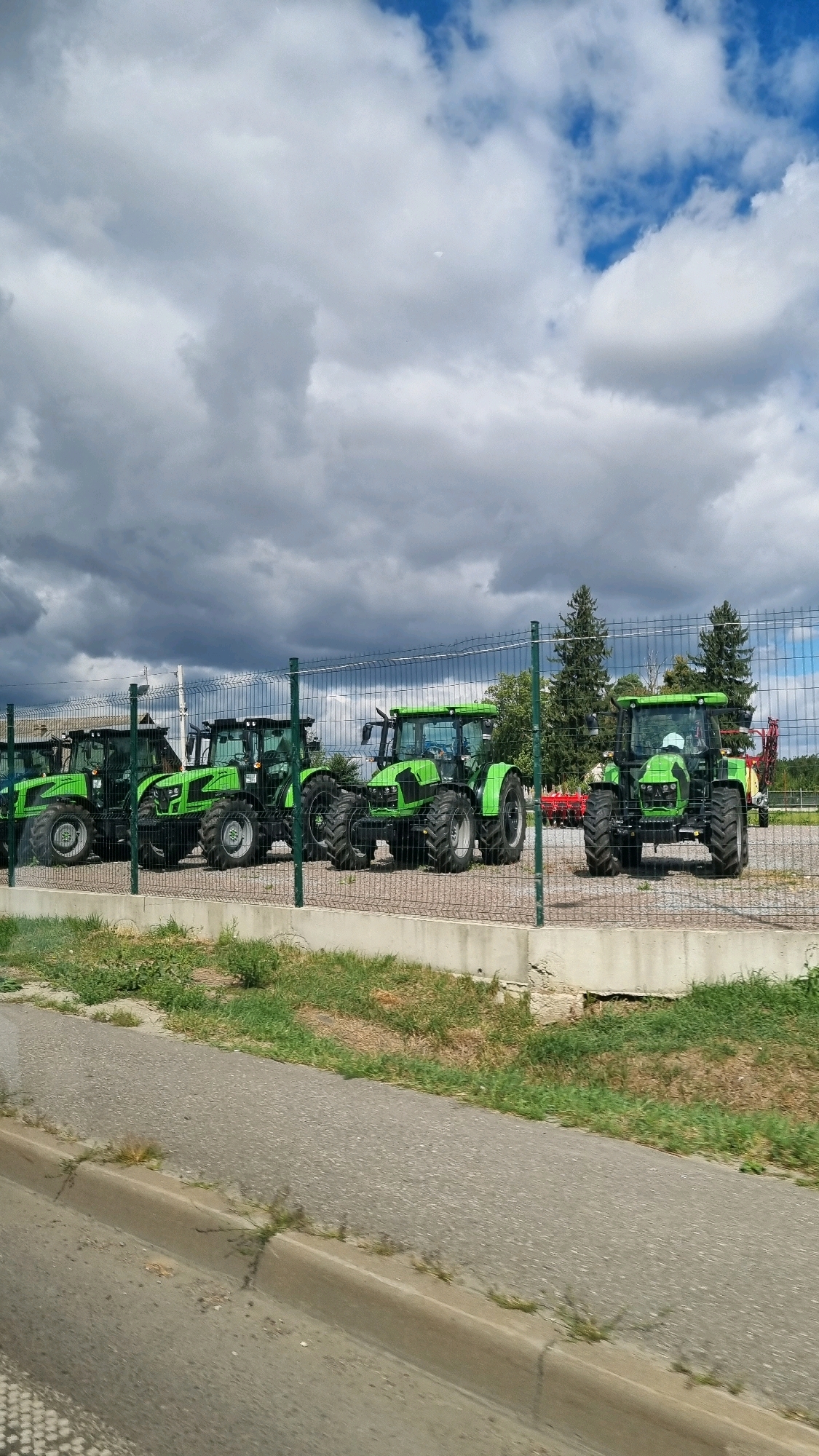 The image shows a lot of green tractors parked in a row behind a metal fence. The tractors are all new and shiny, and they are parked in a large, open lot. The sky is blue with some white clouds. There are some trees in the background. The location is in the city of Kizliv, Ukraine. The scene is a sunny day, and there are no people visible in the image.