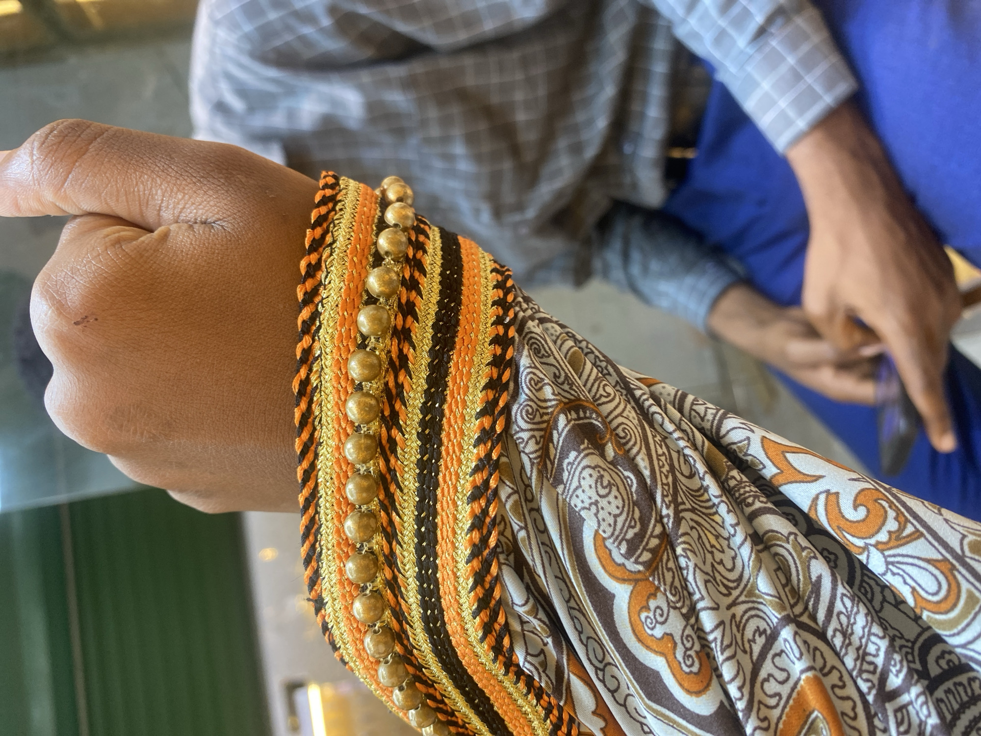 A close-up shot shows a person's arm and hand, possibly indoors in Dar es-Salaam, Tanzania, judging by the style of the patterned sleeve. The arm is adorned with a decorative sleeve that features alternating black, orange, and gold braided bands, accented by a line of gold-colored beads. The person's hand is clenched into a loose fist. In the background, another individual can be seen wearing a gray and blue checkered shirt and blue pants, holding a phone. The scene appears to be taken at a low angle with a green table surface in the lower-left corner.