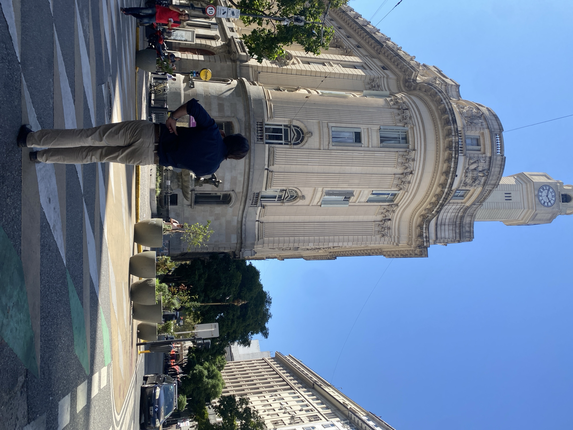 The image shows a street scene in Buenos Aires, Argentina, featuring a man standing on a crosswalk looking at the Palacio de la Legislatura de la Ciudad de Buenos Aires, a government building. The man is wearing a dark blue shirt and khaki pants. The building is tall, ornately decorated with columns and sculptures, and includes a clock tower. The sky is clear and blue. The street includes trees, cars, and several planters.