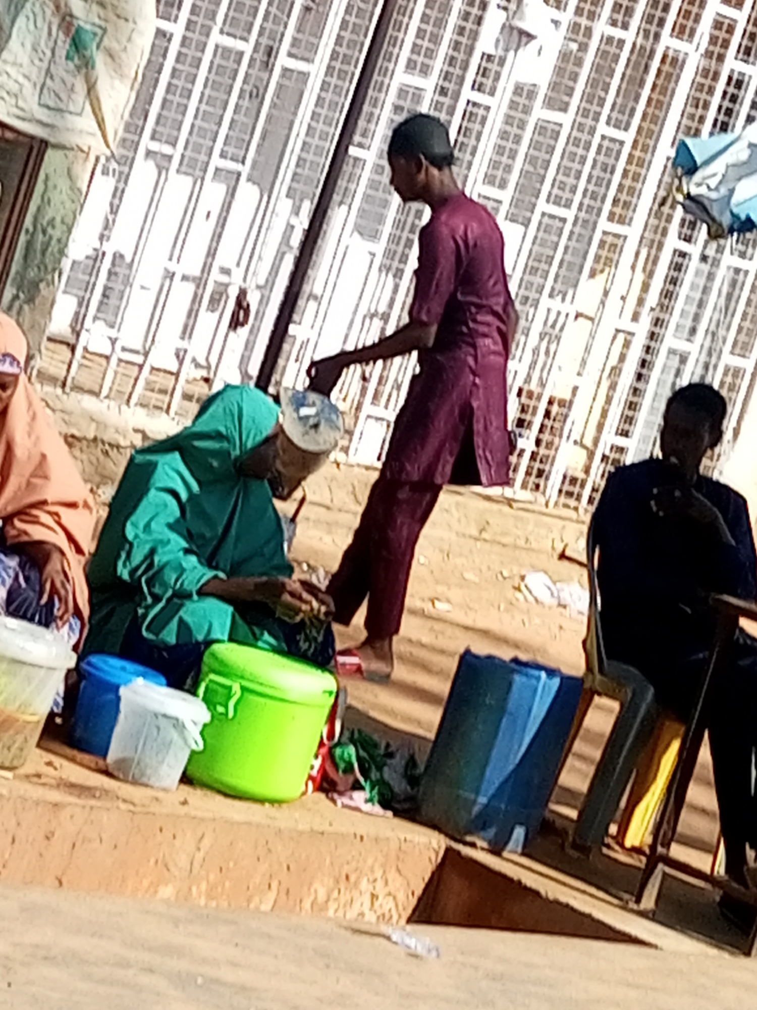 In the city of Tempure, Nigeria, on a sunlit outdoor street, three individuals are present. A young male, wearing a maroon tunic and cap, stands facing left, his right hand near his hip. To his left, a person in a green headscarf and tunic is seated, appearing to be engaged in an activity with their hands. Further left, another individual is seated, covered in an orange garment. In the foreground, several containers are visible: a green bucket, a blue bucket, and a white bucket, along with a larger white bag. The background features a building with a white, patterned facade. The ground is composed of sandy earth.