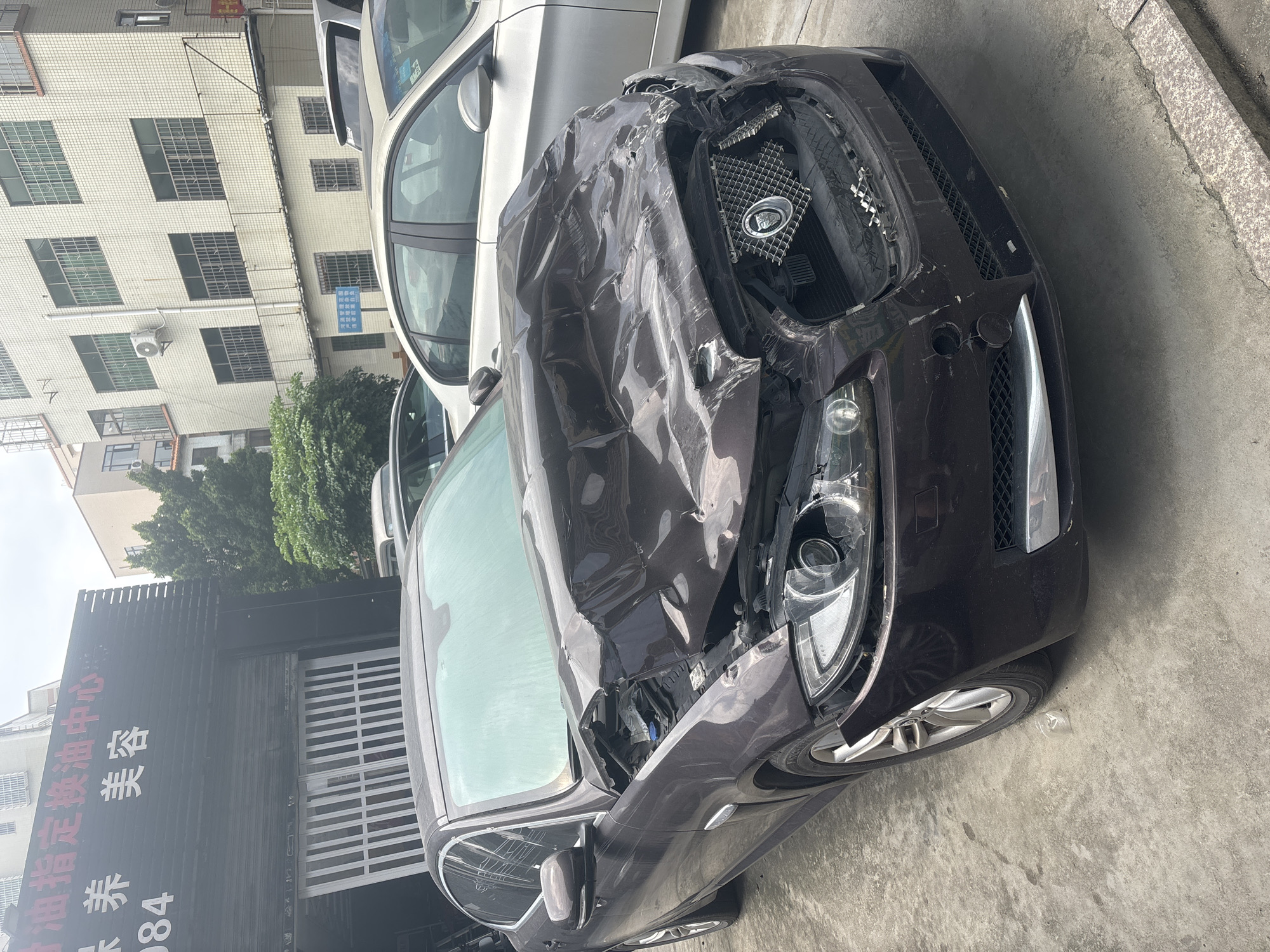 A dark-colored Jaguar sedan, heavily damaged with a crushed hood and front end, is parked on a concrete surface outside a business. The hood is buckled and deformed, obscuring the engine compartment. The grille is damaged, and the headlights appear intact. The car's left front wheel is visible, and a small clear plastic bag lies on the ground near it. Behind the Jaguar, a silver sedan is parked. In the background, a building with multiple windows and air conditioning units is visible, along with trees and an outdoor shop sign with Chinese characters. The scene appears to be in Nanhai District, China.