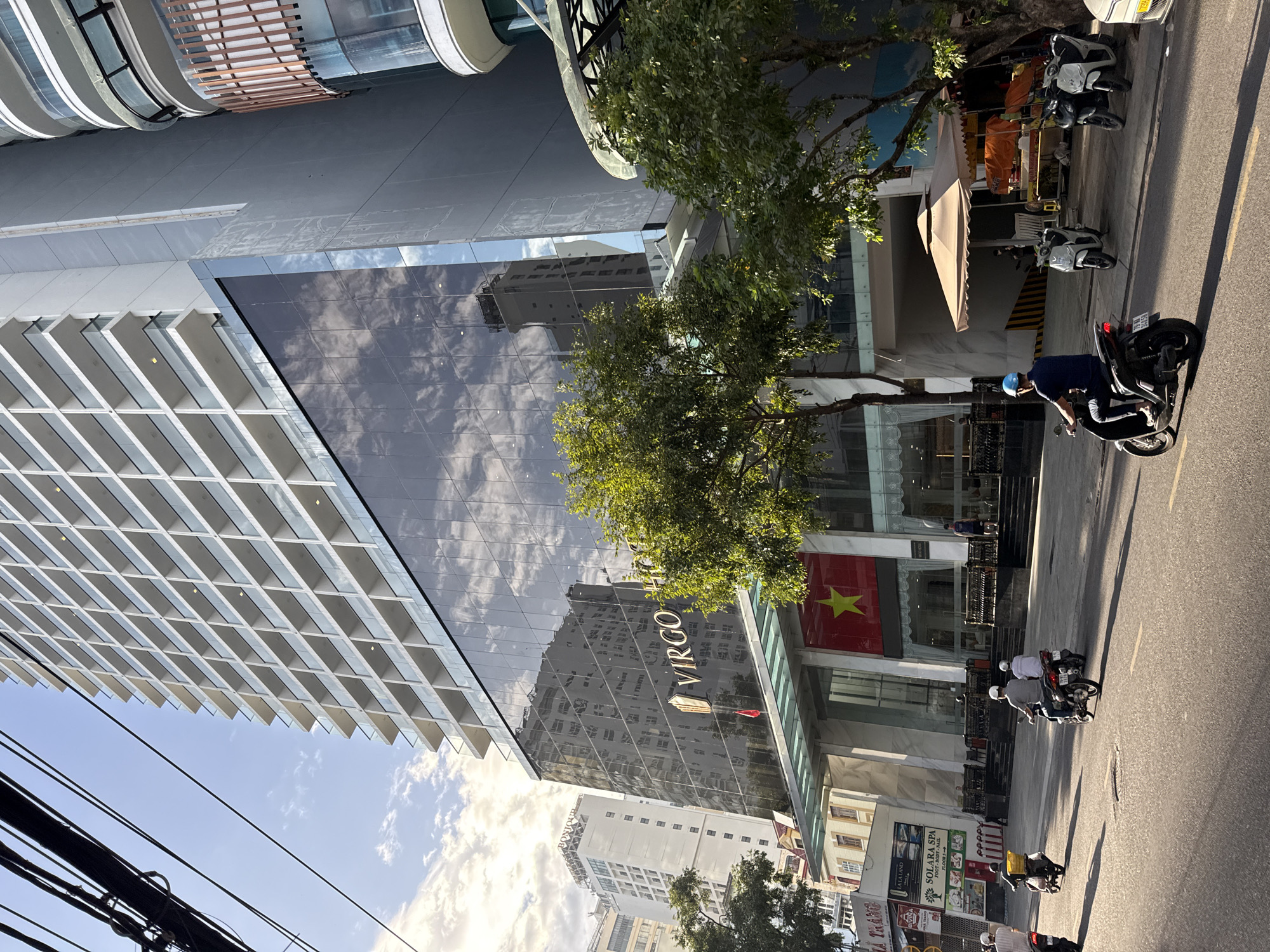 The image captures a street scene in Nha Trang, Vietnam, with a prominent modern hotel named "VIRGO HOTEL" dominating the frame. The hotel features a dark, reflective glass facade that mirrors the sky and surrounding buildings. Above the glass, the hotel has multiple floors with white balconies. In front of the hotel entrance, a large Vietnamese flag with a yellow star on a red background is displayed.
The street in front of the hotel is busy with motorbikes. Several individuals are seen riding motorbikes, some with passengers. One person is wearing a blue helmet. In the foreground, the road is asphalt, and shadows are cast by the sun, indicating it is daytime.
To the left of the hotel, other buildings are visible, including a taller, lighter-colored building with many windows, and a building with signage that includes "SOLARA SPA". Trees with green foliage line the street. On the right side of the hotel, there is a covered seating area with tables and some sort of outdoor stalls. Electrical wires are visible in the upper left corner of the image, crossing over the scene. The sky is partly cloudy with a mix of blue sky and white clouds.