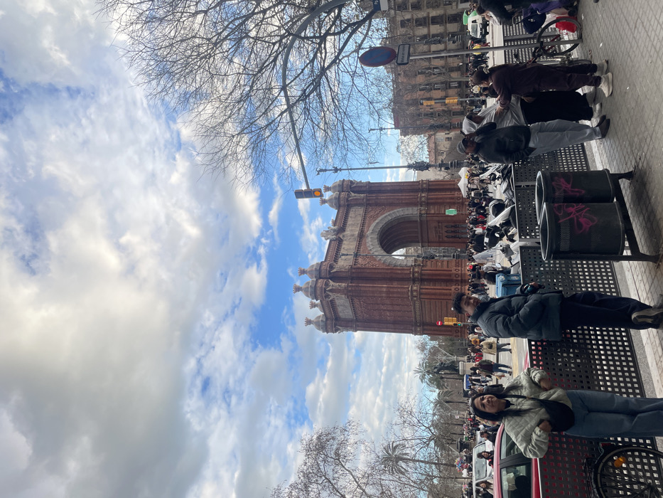 The image depicts a vibrant daytime street scene, with the prominent Arc de Triomf of Barcelona serving as a central architectural feature. The photo is originally rotated 90 degrees counter-clockwise; when viewed correctly, the triumphal arch stands grandly in the midground, with a bustling urban environment surrounding it.
The sky in the upper left portion of the image is a mix of fluffy white clouds and patches of bright blue, suggesting a partly cloudy day. To the right of the sky, bare tree branches extend across the top, indicating late autumn or winter. Below these branches and to the left of the arch, a traffic light is visible, displaying a yellow or orange signal.
The Arc de Triomf is a reddish-brown brick structure, ornately decorated with sculptural details and finials along its top. Its distinctive archway is clearly visible, and various people are gathered in the square or promenade leading up to it. In the distance, beyond the arch, other buildings of an urban landscape can be seen under the bright sky.
In the midground and foreground, the scene is populated with numerous people. On the right side of the image, two men are standing next to a metallic public bench that has two cylindrical trash bins attached. These bins show some pink graffiti. Further into the foreground on the right, two women are seated or leaning on a red object, possibly a car. One woman, with dark hair, is wearing a light-colored quilted jacket and jeans, with a black fanny pack across her chest, and is looking towards the camera. The other woman is partially obscured.
A crowd of people can be observed near the base of the Arc de Triomf, some seemingly walking along the wide promenade, while others appear to be gathered in groups. There are also what look like white tents or stalls set up further down the path towards the arch, suggesting a market or a public event. Several bare-branched trees are scattered throughout the scene, and some palm trees are also visible further in the background. A blue circular sign with a red diagonal line (indicating no stopping or standing) is mounted on a pole above the people on the right side. Below it, a white rectangular sign with black text is present, but the text is not legible. A bicycle is also visible parked near some people on the far right. The overall impression is of a lively public space on a bright day.