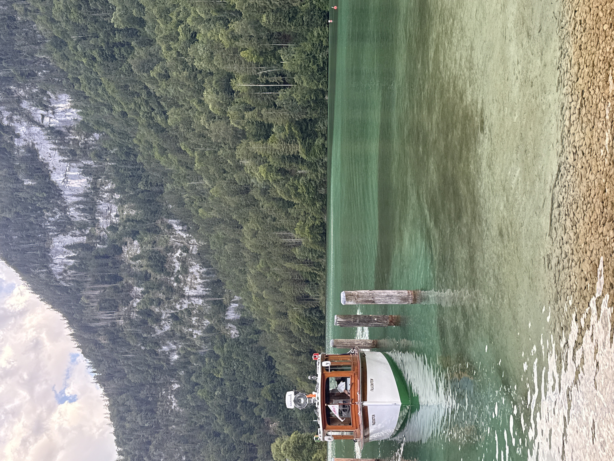 A boat named "FALLENSTEIN" is docked at the Königssee lake in Schönau am Königssee, Germany. The boat is white with a green stripe and a wooden cabin. A person is visible steering the boat. The lake is a clear, turquoise green and surrounded by steep, forested mountains. Wooden pilings are in the water near the boat. The sky is cloudy with patches of blue.