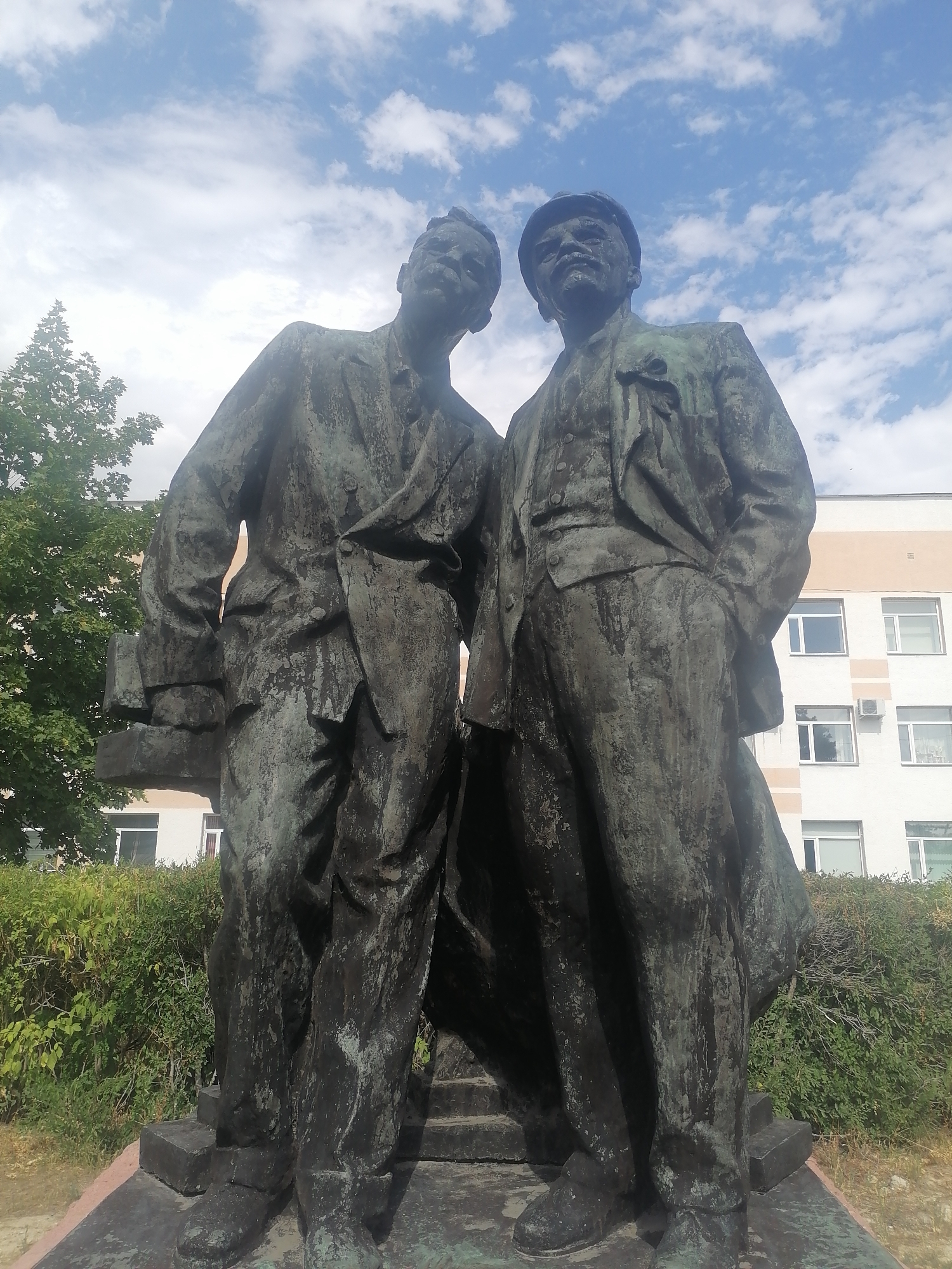 A weathered bronze statue depicts two men in suits standing side-by-side, positioned outdoors in front of a light-colored building with multiple windows. The statue's surface shows signs of age, with a greenish patina and dark streaks. The man on the left is slightly taller, holding what appears to be a book or folder in his left hand. The man on the right wears a cap and has his hands in his pockets. Both figures are depicted in formal attire, with the suits having visible lapels, buttons, and trousers.
Behind the statue, a dense green hedge forms a backdrop, with a large tree on the left side of the frame. The building in the background has a horizontal band of darker color separating its upper and lower sections. The sky above is blue with scattered white clouds. The statue is situated on a raised platform, composed of dark stone blocks.
This statue is located in Karaganda, Kazakhstan, and represents a historical monument.