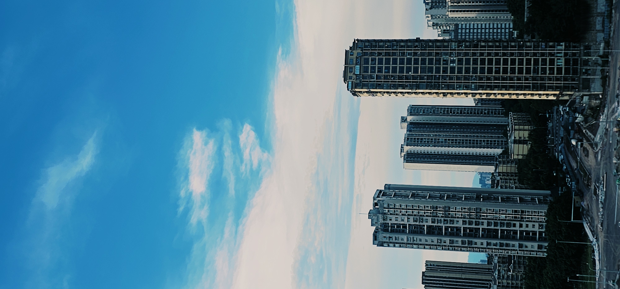 This is a vertical, eye-level medium shot of a city skyline in Jiulongpo District, China, under a bright blue sky with scattered white clouds. The foreground shows a construction site with dirt, gravel, and piles of materials, along with streetlights and a cement mixer. Beyond the construction, a dense urban landscape of numerous tall, modern apartment buildings stretches across the mid-ground. Some buildings are closer and more detailed, while others are further away and appear as a mass of high-rises. A layer of dark green trees is visible in the lower-mid ground, interspersed between the buildings and the construction site. The overall impression is of a rapidly developing urban area.
