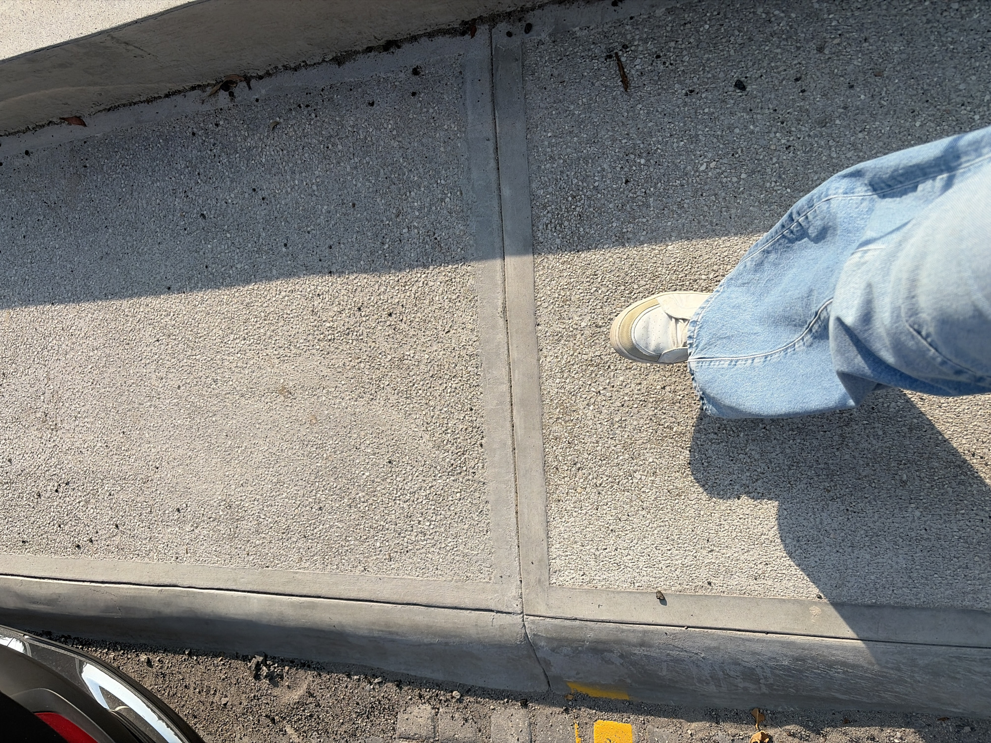 A high-angle shot shows a person wearing light blue jeans and white sneakers standing on a concrete sidewalk in Puebla, Mexico. The sidewalk is segmented into sections with concrete dividers. A car's headlight is visible in the lower left corner. The curb is visible along the lower edge of the sidewalk, and some paving stones are visible below the curb. Yellow paint is on the curb and a dried leaf lies on the curb's edge. The scene is brightly lit.