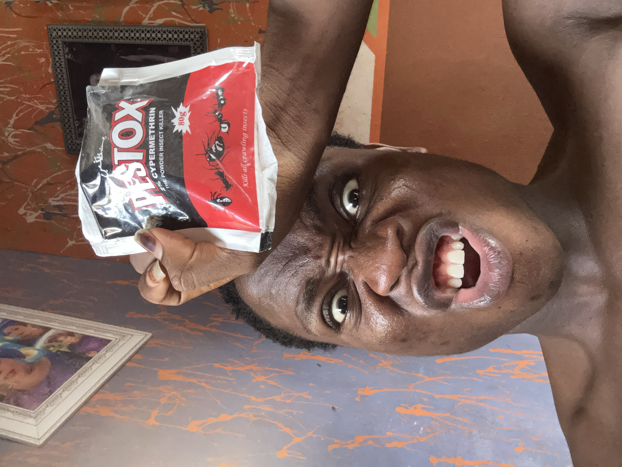 A dark-skinned individual is holding up a packet of Pestox insecticide. The packet is white and red and has the words "PESTOX 2% CYPERMETHRIN THE POWDER INSECT KILLER" written on it. The individual is making a face, with their eyes wide and mouth open. The background is a room with orange and grey walls. There is a framed picture on the wall behind the individual. Another framed picture is partially visible on a table or other surface in the lower left corner of the image. The image is taken from a low angle, making the individual appear to be looking down at the viewer. The location is Unknown, Unknown.