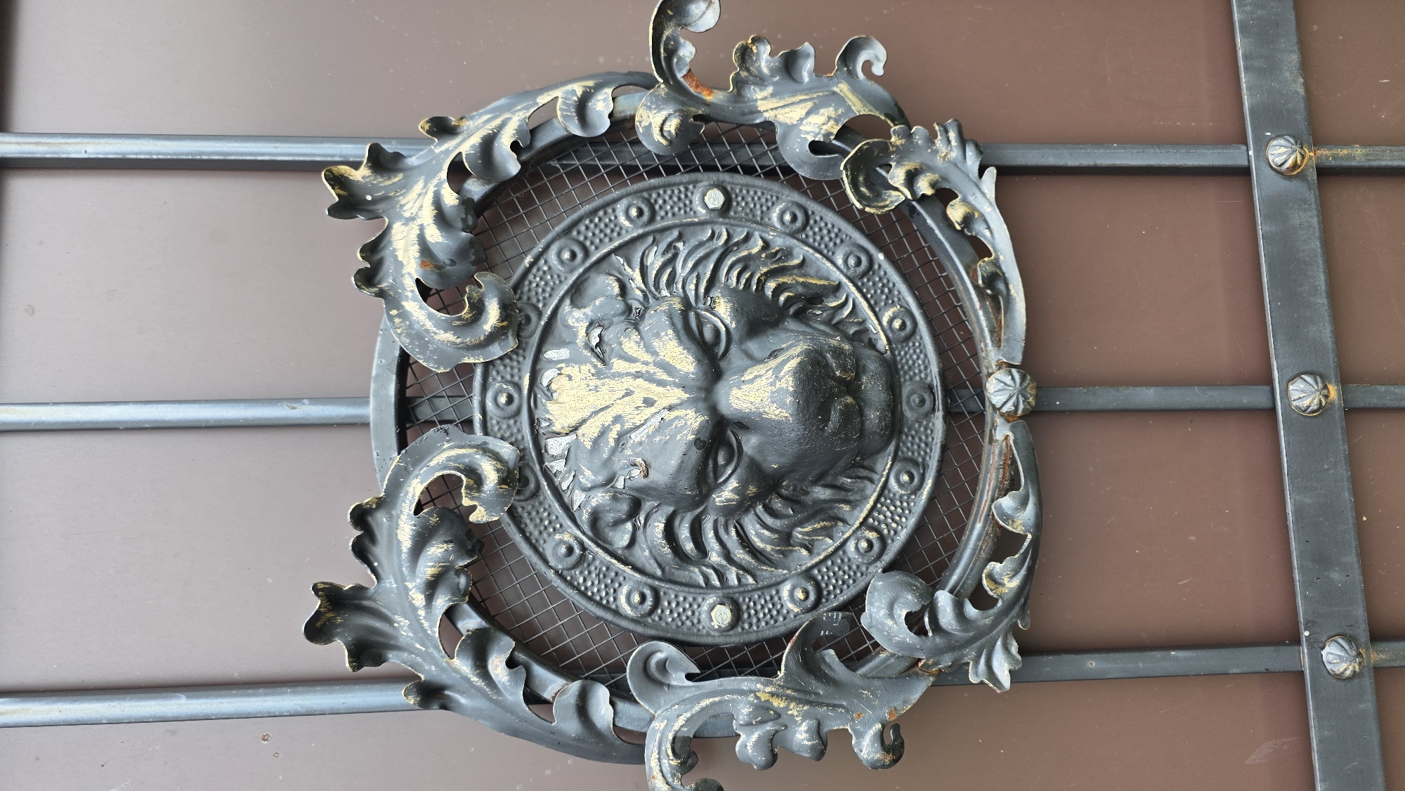 The image displays a close-up, eye-level view of an ornate, dark metal decorative element on a gate in the village of Starosharyevo, Russia. This element features a central, detailed bas-relief of a lion's head, with a textured mane and expressive eyes. The lion's head is encircled by a circular frame adorned with raised dots and concentric rings. Extending outwards from this central circle are decorative scrolled leaf-like metalwork pieces, some of which are accented with a distressed gold paint effect, showing wear and age. The entire decorative piece is mounted on a series of vertical and horizontal dark metal bars, part of a larger gate structure. The background is a flat, matte brown surface, likely the gate itself, with some subtle variations in color and light. The image is sharp and well-lit, highlighting the intricate details of the metalwork. There are no people or animals visible in the frame. The context of Starosharyevo, Russia, suggests this is an outdoor setting, possibly at the entrance to a property.