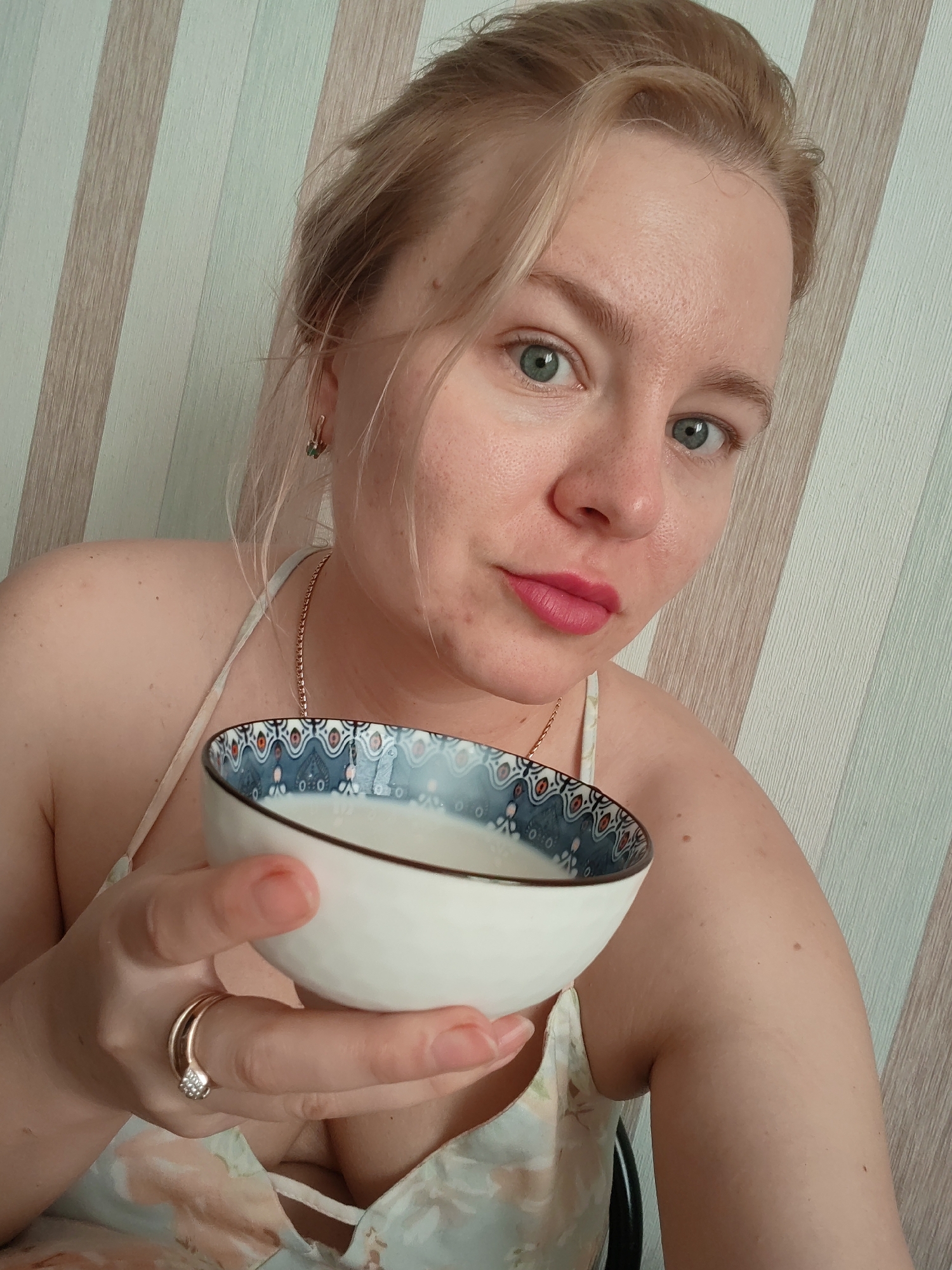 A medium shot shows a fair-skinned woman with blonde hair, green eyes, and pink lipstick holding a white bowl with a patterned blue and white rim. She is wearing a light-colored camisole with a floral pattern and a gold chain necklace. A gold ring with small stones is on her right ring finger. The background consists of vertical stripes in shades of beige and white. The scene appears to be indoors, potentially in a residential setting, and there is no indication of the city of Izhevsk, Russia.