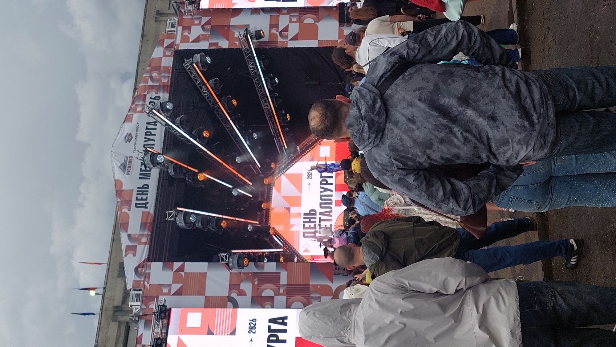 This is a medium shot image captured outdoors, looking towards a stage setup under an overcast sky. The stage has a large backdrop with "ДЕНЬ МЕТАЛЛУРГА 2026" (Metallurgist's Day 2026) prominently displayed, along with logos for "РУСОВО" and "ГЕОПРОМИНВЕСТ". The stage is equipped with multiple lighting rigs, some emitting orange and white lights. A performer in a blue outfit is visible on stage. In the foreground, a crowd of people are gathered, facing the stage. Several individuals in the foreground are wearing hooded jackets and casual clothing, with a focus on their backs. The ground appears to be paved or damp. The overall scene suggests an outdoor event or festival.