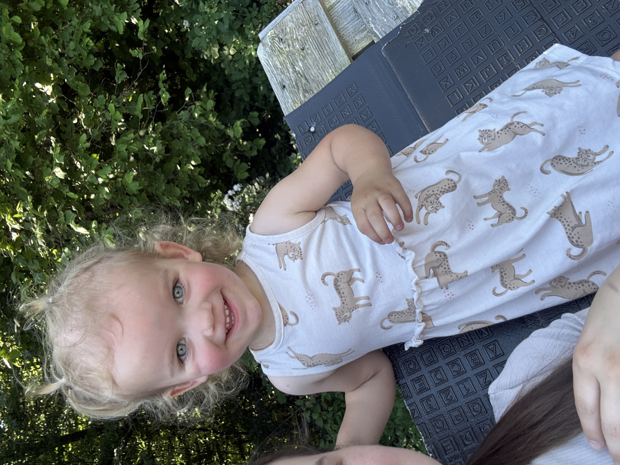 A young child with blonde curly hair styled in two pigtails is smiling and looking at the camera. The child is wearing a white sleeveless dress with a repeating pattern of leopards and pink dots. They are sitting on a dark textured surface, possibly a bench or mat, next to a wooden structure. In the background, there are green leafy trees and some white flowers. Part of another person's shoulder and hair is visible on the left side of the frame. The scene appears to be outdoors in a garden or park setting. The image was taken in Beltershain, Germany.