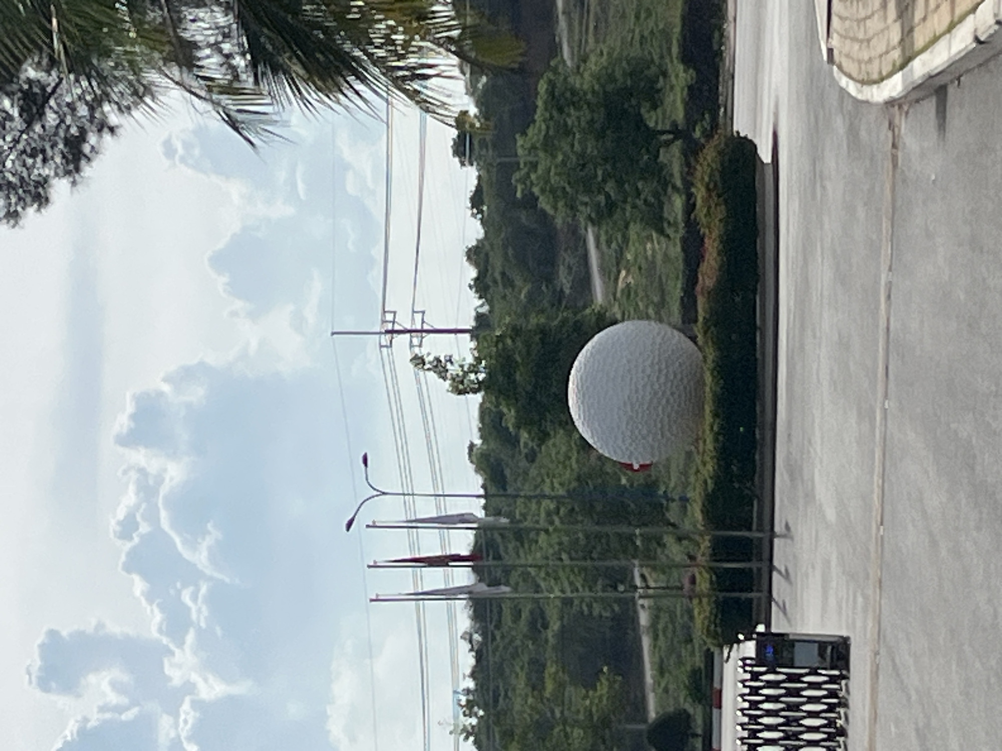 The image depicts a street scene in Phu Thuy Ward, Vietnam. The foreground is dominated by a large, white, dimpled sphere, resembling a golf ball, positioned on a landscaped area. Behind the golf ball, a row of flags on poles are visible, with a double-headed lamppost above them. Further in the background, lush green trees and foliage form a dense backdrop, interspersed with power lines and utility poles. A paved road or pathway runs alongside the landscaped area. The sky is partly cloudy with large, fluffy white clouds against a light blue sky. The lighting suggests it is daytime, likely mid-morning or afternoon, with overcast conditions. The scene appears to be an outdoor public space, possibly a park, golf course, or roadside attraction. No people are visible in the image.