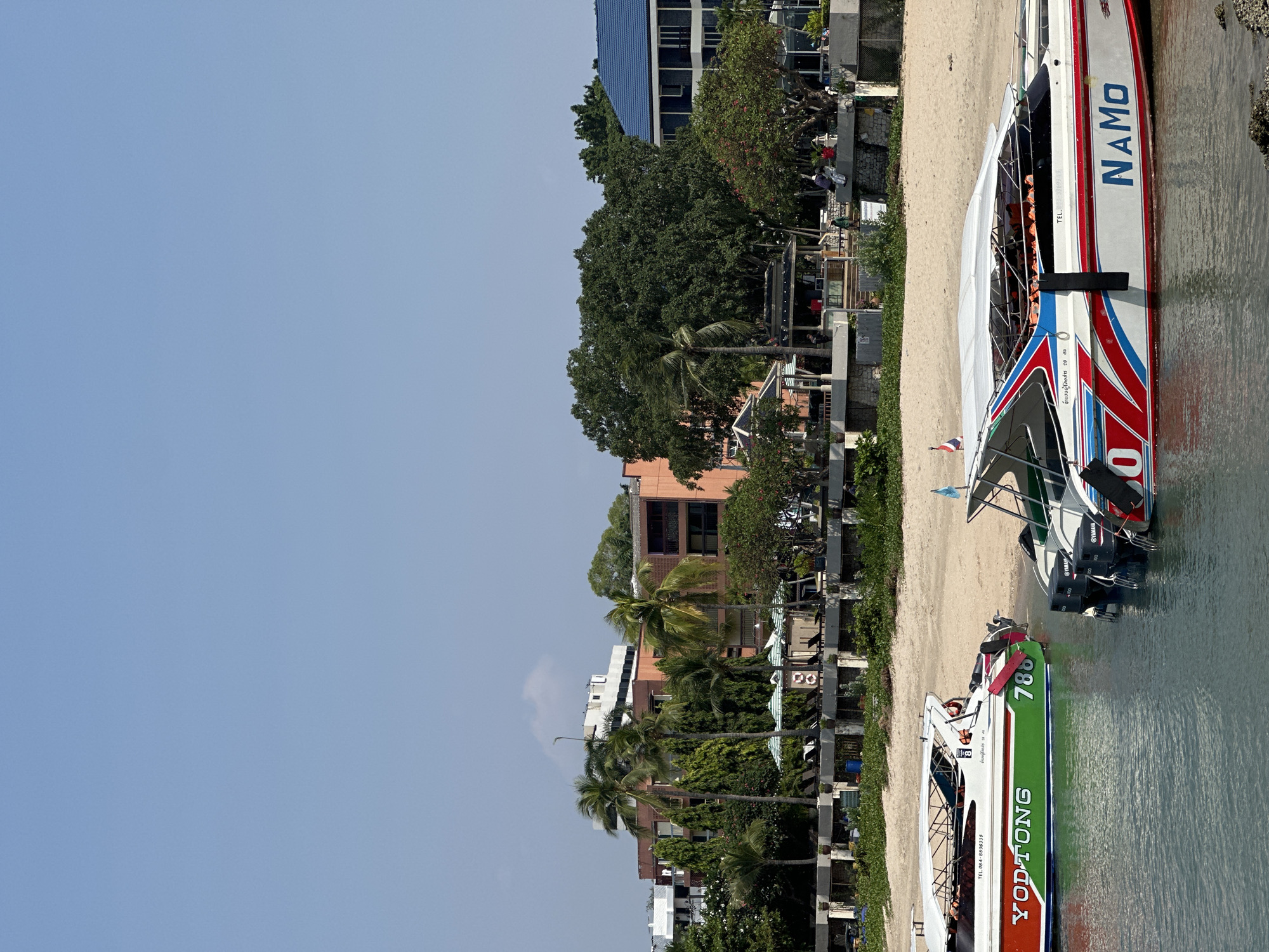 This image shows a view of a beach in Pattaya City, Thailand. The image is taken horizontally and then rotated 90 degrees counter-clockwise. The beach is lined with buildings and trees. There are two speedboats in the foreground, which are partially in the water. The boat closer to the viewer is labeled "YODTONG" and the other is labeled "NAMO". The sky is clear and blue.