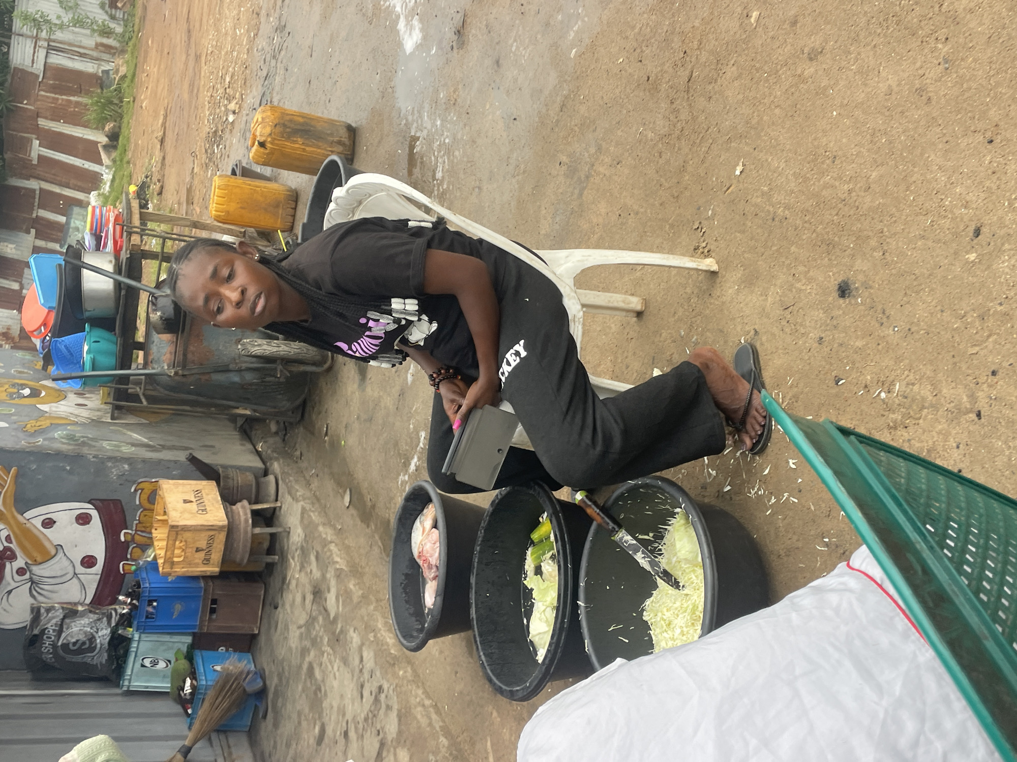The image depicts a woman sitting on a white plastic chair next to three large black basins in what appears to be an outdoor market area. The woman is wearing a black t-shirt and black pants, and she is holding a tablet. The first basin contains raw fish, the second contains chopped cabbage and cucumbers, and the third contains shredded cabbage, with a knife lying in it. Behind the woman, there are two large yellow plastic containers and a metal cart with various kitchenware. In the background, there are stacks of plastic crates, a broom, and a mural painted on a building. The ground is a dirt surface, appearing wet in some areas. The scene is likely in Jos, Nigeria, as indicated by the presence of the woman's attire and the general setting, which is characteristic of market environments in Nigerian cities.