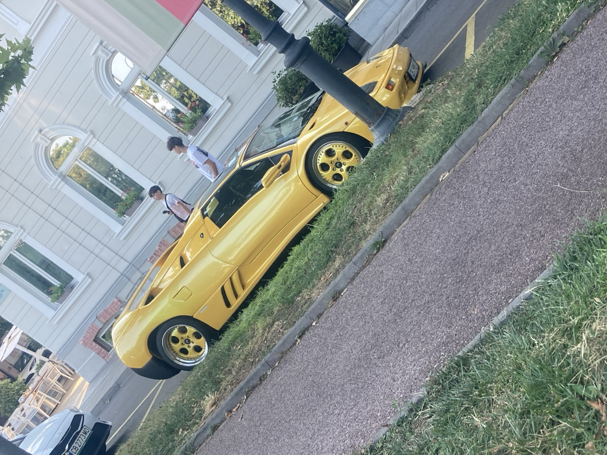 A bright yellow Lamborghini Diablo is parked on a grassy verge next to a paved walkway. The car has gold rims and a black spoiler. Two young men, one wearing a white t-shirt and the other a striped t-shirt, are standing near the car. In the background, there is a light-colored building with arched windows, and outdoor cafe seating. To the left of the Lamborghini, a white car with a license plate starting with "CB 2773 MO" is visible. A black lamppost is in the foreground on the right side of the image. This scene is located on a street in Burgas, Bulgaria, likely in the area of the Sea Garden, given the architecture and presence of outdoor dining.