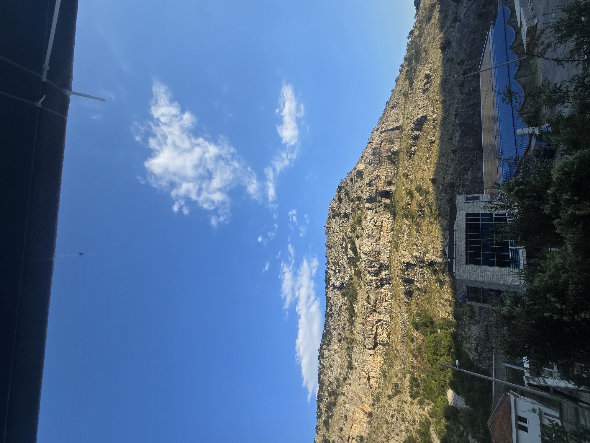 The image depicts a wide shot of a mountainous landscape under a clear blue sky. A sheer, rocky mountain face dominates the background, its rugged terrain casting shadows. In the middle ground, several modern buildings are visible. One building is constructed with stone and glass, featuring large windows. Adjacent to it is another structure with a blue roof, possibly a greenhouse or industrial building, partially covered with solar panels. Greenery, including trees and bushes, dots the landscape, interspersed with the buildings. In the foreground, a dark overhang obstructs the very top of the frame, from which a thin string hangs. The overall scene suggests an outdoor setting, likely on the outskirts of a settlement, given the juxtaposition of natural mountain scenery and constructed buildings. The lighting indicates it is daytime.