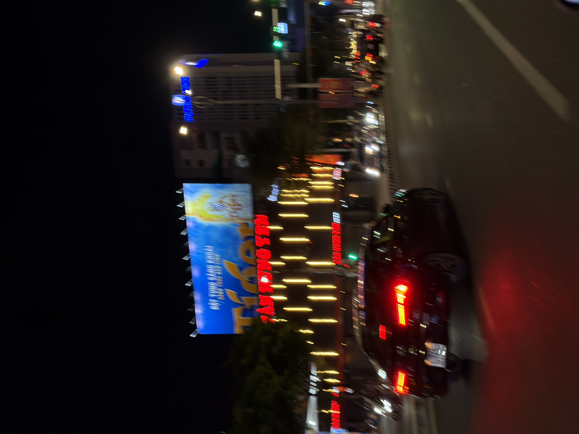 The image captures a street scene at night in Da Nang, Vietnam, likely on a thoroughfare with commercial establishments. A prominent billboard advertising "Tiger" beer, featuring a tiger graphic and Vietnamese text including "ĐÁT TUNG SẢNG KHOÁI", is visible in the upper portion of the frame. Below the billboard, a building with glowing vertical yellow lights and red neon signs that read "TÂN HUNG YÊN" are illuminated.
In the foreground, a dark-colored car with its taillights illuminated in red is driving away from the camera. The car's license plate is partially visible and appears to be obscured by motion blur. Multiple other vehicles, including cars and motorbikes, are also present on the road, with their headlights and taillights creating streaks of light due to the motion. The overall scene depicts a busy urban environment at night, characterized by artificial lighting from billboards, signs, and vehicles. The motion blur suggests the photograph was taken from a moving vehicle.