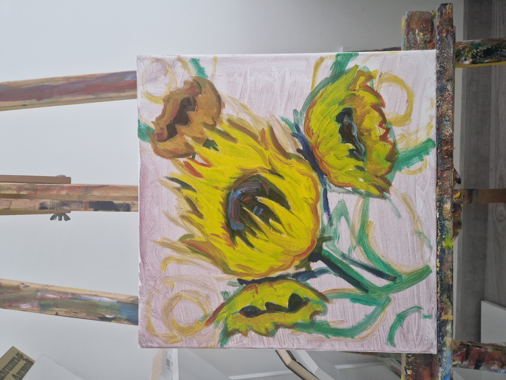 The image shows an oil painting of sunflowers on a canvas, positioned on an easel indoors. The painting features three main sunflowers with vibrant yellow petals and darker centers, accompanied by green stems and leaves. The background is a muted pink or off-white. The easel is made of wood and shows splatters and markings from previous painting projects. The setting appears to be an artist's studio, with a plain white wall visible in the background. The location in the city of Unknown, Unknown is unknown.