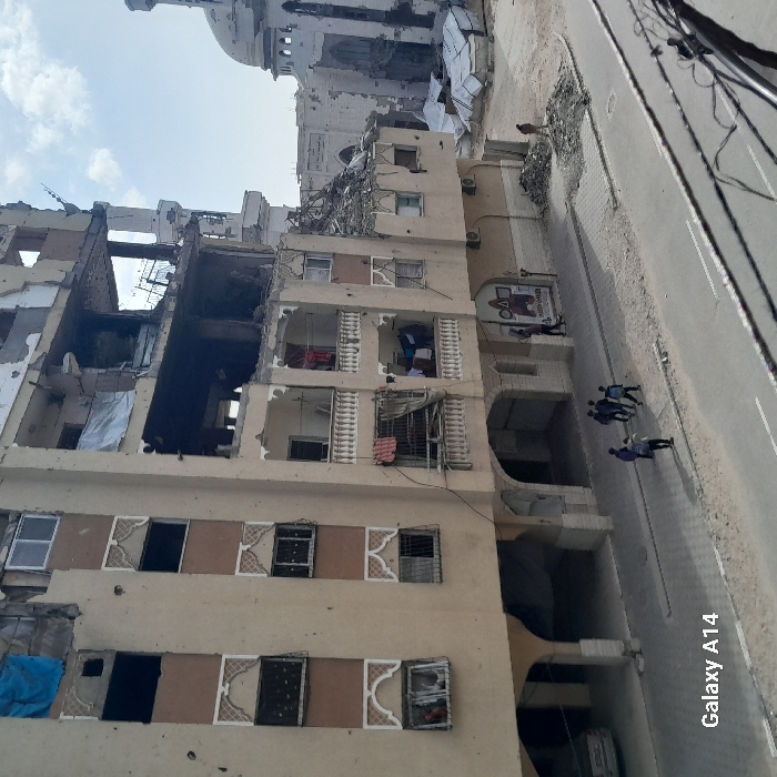 A damaged building stands beside a road with several people walking on it. Part of the building is missing, revealing inner rooms. Adjacent to the damaged structure, a more intact beige building features decorative window frames. A billboard hangs on the undamaged building. Overhead, a white domed structure is visible, suggesting a mosque or significant religious building. The scene implies war or conflict.