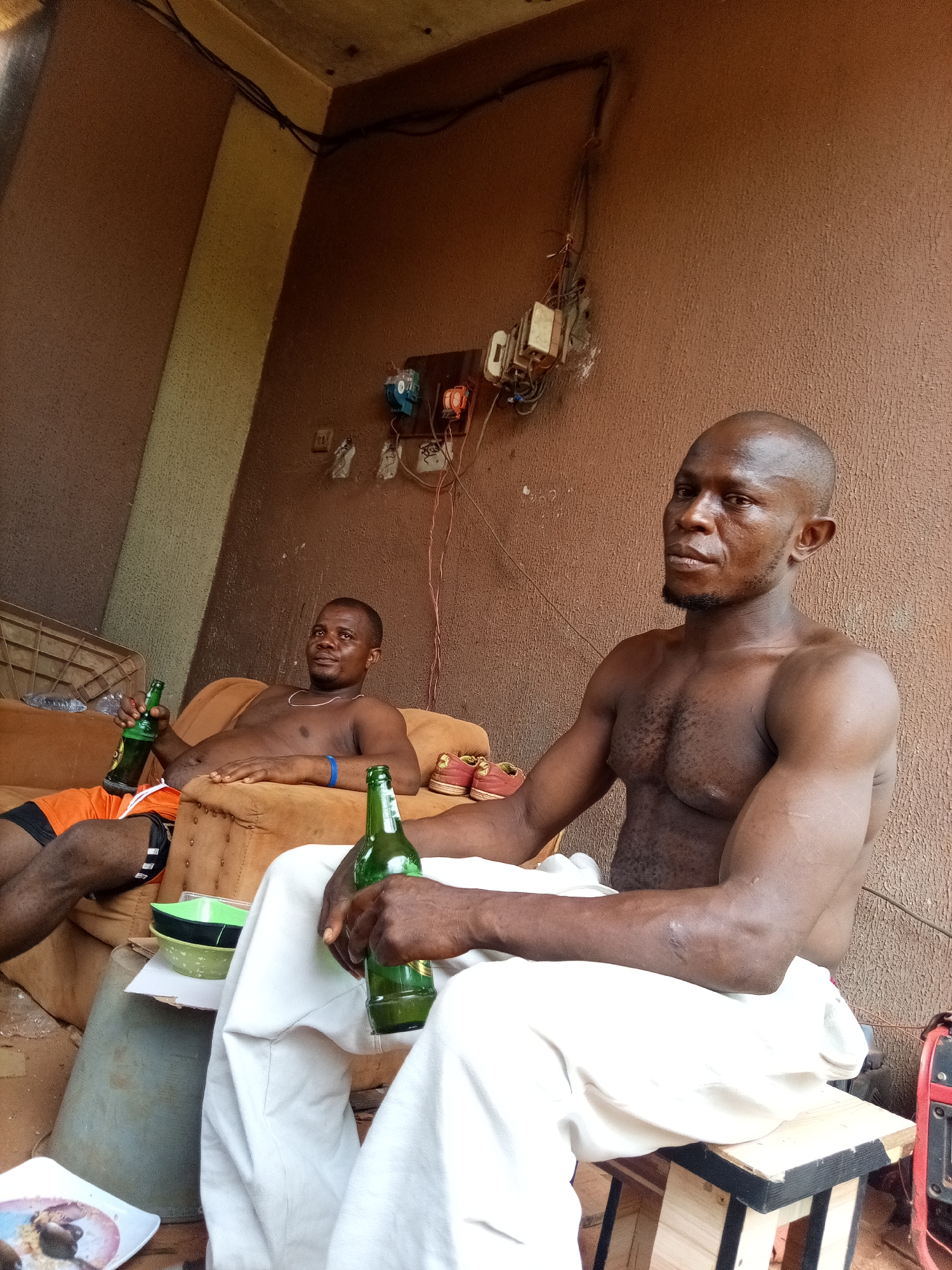 Two shirtless African men are seen outside a building in Ukwu-Abwa, Nigeria. The man in the foreground is sitting on a wooden stool covered with white cloth, holding a green beer bottle. He has a shaved head and a beard. The second man is reclining on a brown sofa in the background, also holding a green beer bottle. There's an electrical panel on the brown wall behind them. A table with bowls, a bucket, and a plate of food are situated in front of the man on the stool. A red generator is visible to the right of the man on the stool.