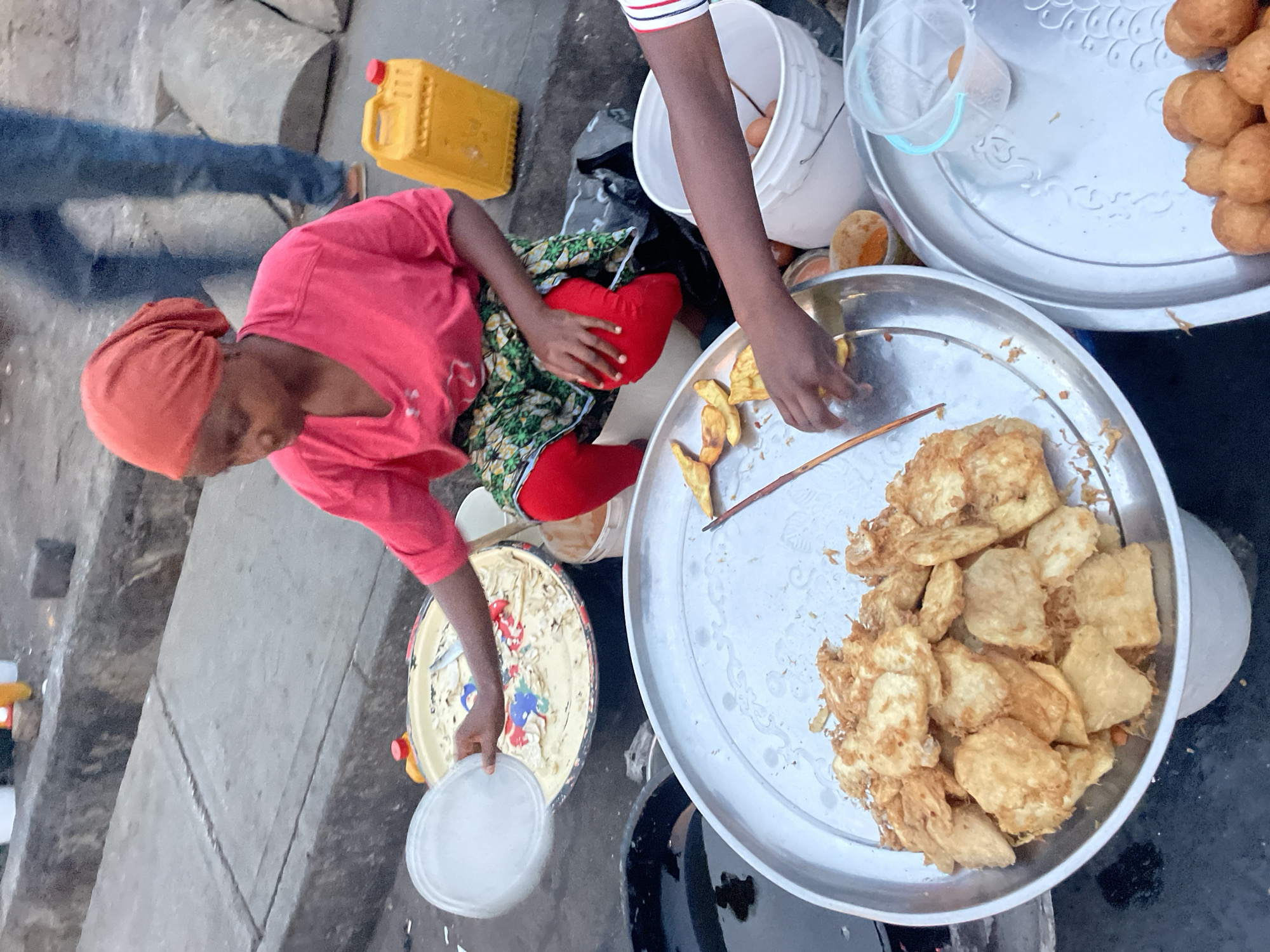 A woman in a red top and headscarf sits near a street curb, selling food. She is positioned next to an arrangement of food preparation and sales items, including metal trays with fried food, a yellow jerrycan, and plastic containers. Another person, wearing a striped shirt, is seen reaching towards one of the food trays. The items and action suggest a food vendor or market scene. The location of Jos, Nigeria, is not evident within the visual elements of the image.