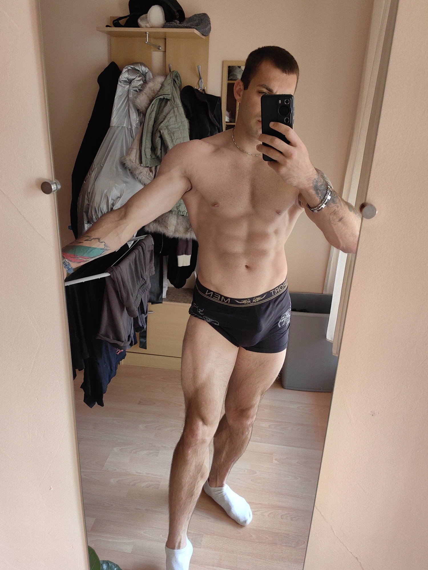 A shirtless person with visible abdominal muscles and tattoos on their left arm is taking a mirror selfie in a room that appears to be a bedroom or dressing area. The person is wearing black underwear and white socks. In the background, there is a closet with several coats hanging on hangers, a shelf with hats and other items, and a trash can. The room has light-colored walls and wooden flooring. The reflection in the mirror also shows a door with a round handle on the left side of the frame. The scene is indoors and appears to be a private residence. There is no discernible connection to Varna, Bulgaria, in the image itself.
