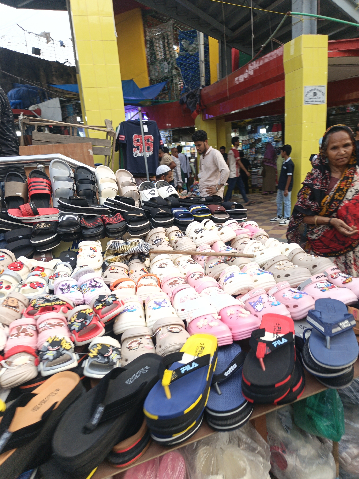 This image captures a bustling market scene, likely a footwear stall, during the daytime. The foreground is filled with a large, diverse display of shoes and sandals, arranged on tables or shelves.
Numerous pairs of adult sandals are visible, including various flip-flops and slip-on styles in colors like black, blue, red, and beige. Some of these feature brand names such as "NIKE," "FILA," "Lee," and "totto," with visible sizes like "40" and "41." Prominently displayed are rows of children's clogs, styled similarly to Crocs, in pastel shades of pink, beige, and white. Many of these clogs are decorated with cartoon characters, such as Batman, and other whimsical designs; some have the text "HEYUN" printed on them.
Several people are present, indicating a lively market atmosphere. In the right foreground, an older woman wearing a patterned headscarf and dress looks towards the camera with a neutral expression. Behind the shoe display, a young man in a light-colored shirt stands, possibly attending the stall, with another man in a white skullcap partially visible beside him. Other individuals are blurred in the background, moving or standing.
Overhead, bright yellow pillars and red beams frame the market structure, suggesting a covered or semi-covered outdoor setting. A dark blue T-shirt with "86 Chicago" printed on it hangs above the shoe display in the mid-ground. The natural light suggests it is daytime. The scene depicts a common market activity of buying and selling footwear.