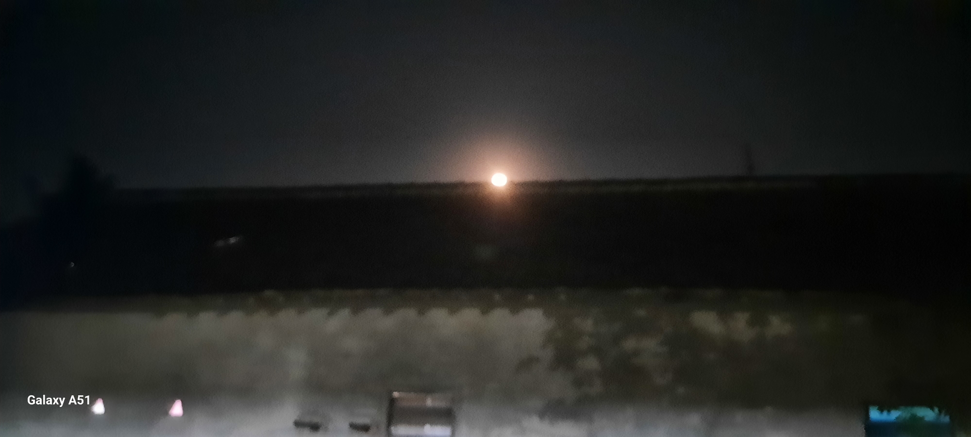 A nighttime exterior shot depicts the moon illuminating the sky above a building. The moon, a bright, circular orb, is positioned in the upper center of the frame. Below the moon, a dark, horizontal structure, likely the roofline of a building, extends across the image. The lower portion of the image shows a textured, lighter-colored wall, possibly stucco or concrete, with a wavy, scalloped edge along the top. A small, rectangular opening, possibly a window or vent, is visible on the wall. The Samsung Galaxy A51 logo is present in the bottom left corner. The scene appears to be located in Ploso, Indonesia, suggested by the branding. The overall atmosphere is dark and serene, with the moon as the primary light source.