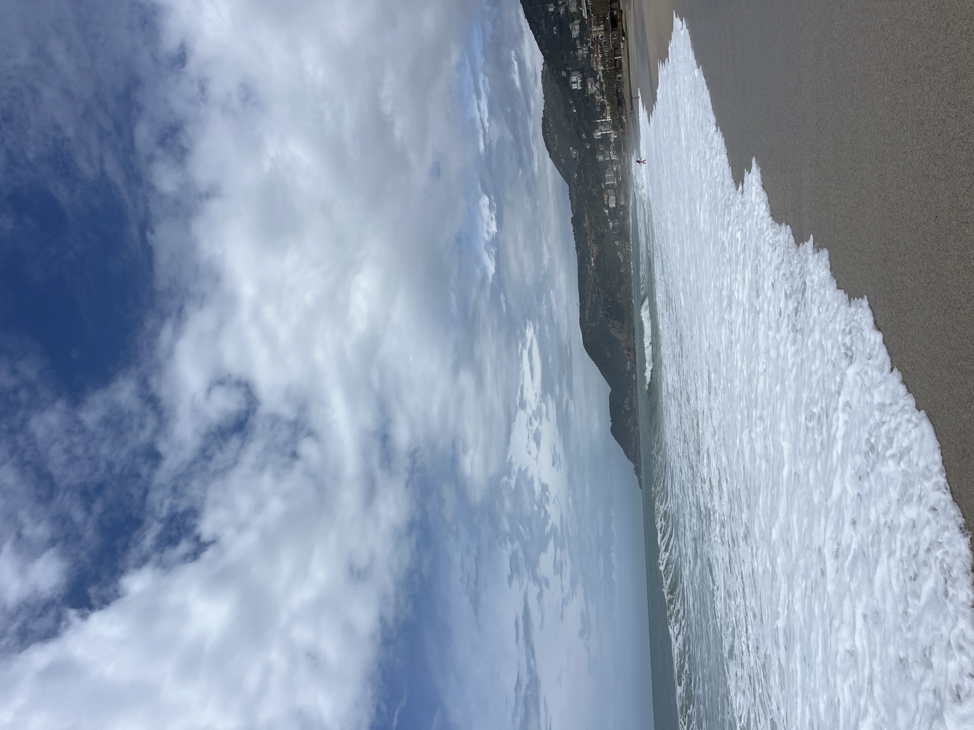 The image depicts a beach scene in Alanya, Turkey, characterized by a sandy shore meeting the sea. A mountain range is visible in the background, hosting buildings and settlements. The sky is filled with clouds, suggesting a partly cloudy day. Waves are breaking on the shore, generating foam, and there appears to be a person in the distance near the shoreline.