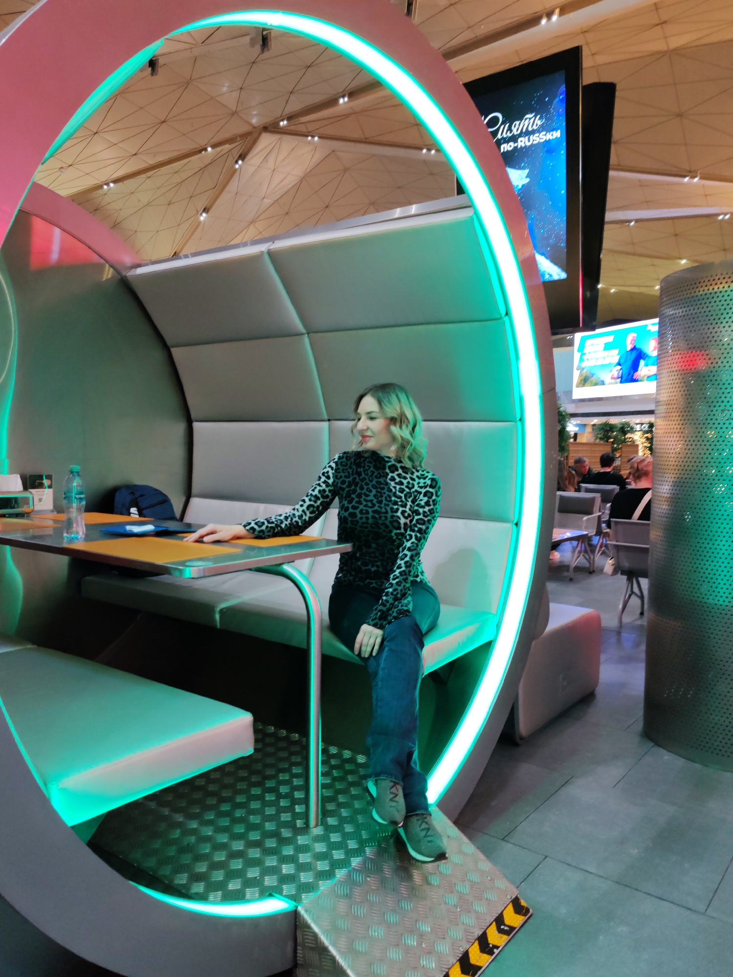 The image depicts a woman sitting in a modern seating pod at Pulkovo Airport in Saint Petersburg, Russia. The pod features a curved frame illuminated with green LED lights and a pink accent on the outer edge. The interior has cushioned seating and a small table. A water bottle and some papers are on the table. The woman is wearing a black and leopard print top, blue jeans, and green sneakers. In the background, there is a large screen displaying Russian text, another screen showing a man in a blue suit, and other people sitting at tables. The ceiling is a complex geometric pattern. A metal, perforated cylinder stands to the right of the seating pod.