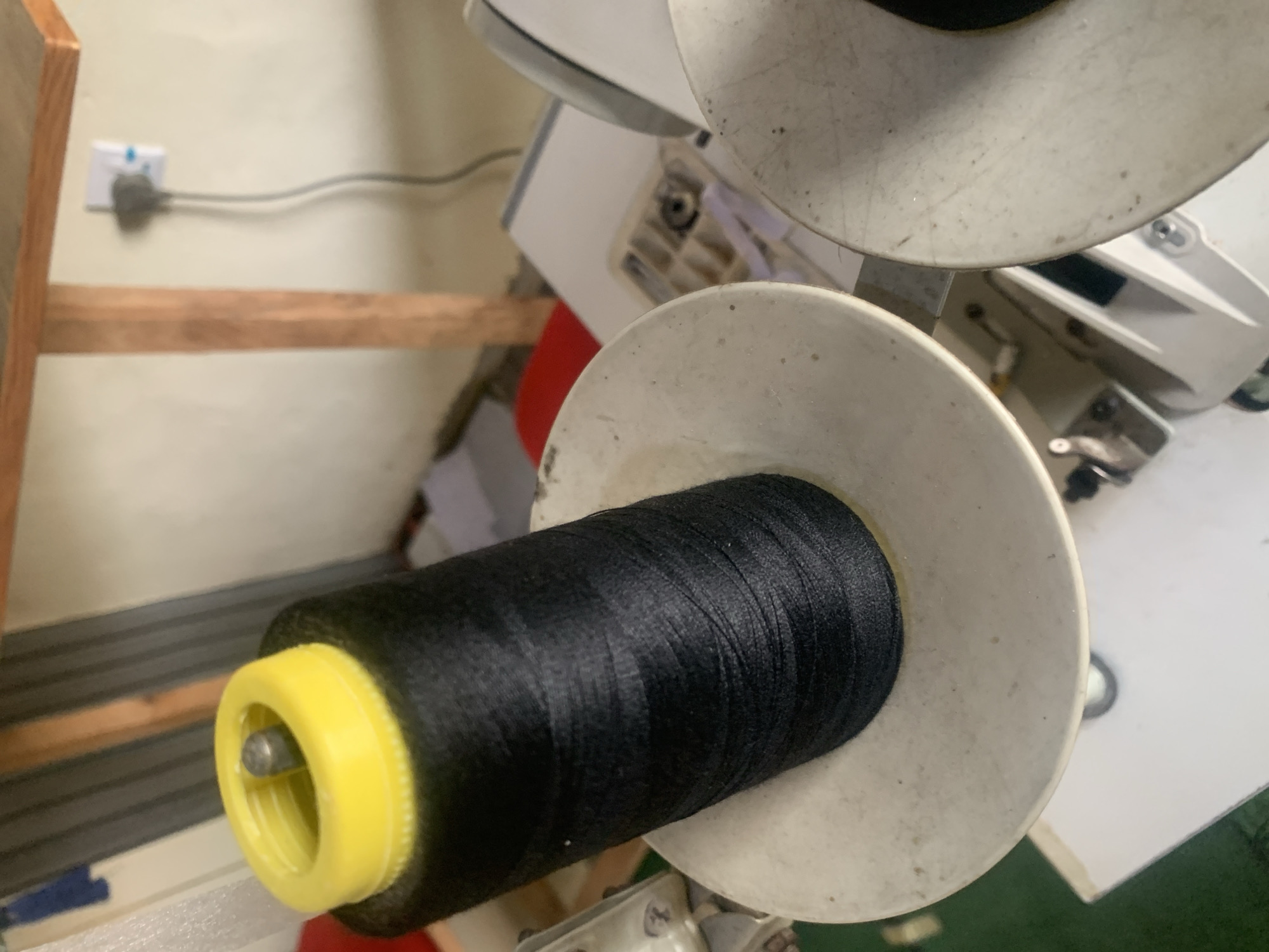 A close-up shot captures a large spool of black thread with a yellow plastic top, positioned on a sewing machine. The spool sits on a white platform, likely part of the machine. In the background, a light-colored wall is visible, with a wooden support beam and a white electrical outlet with a plug attached. The setting appears to be an indoor workshop or domestic space in Jauro Yinu, Nigeria, possibly a tailoring shop or a room where sewing activities take place. The overall impression is one of a functional workspace, with the focus clearly on the sewing thread and machine.