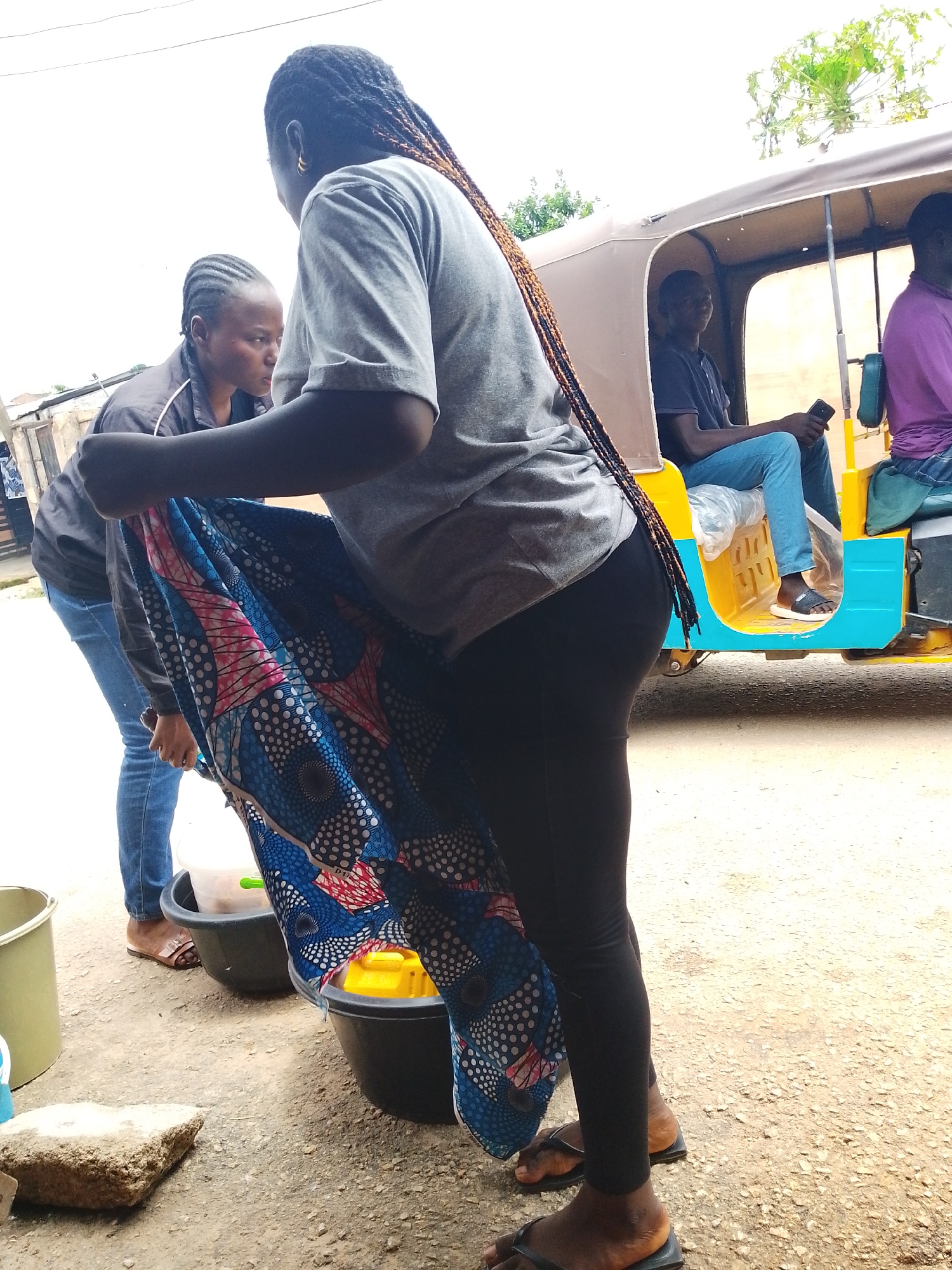 Two women are washing fabric with soap in black buckets. They are outside, on a dirt road. Behind them is a blue and yellow commercial tricycle taxi with two passengers. The setting is Jos, Nigeria.