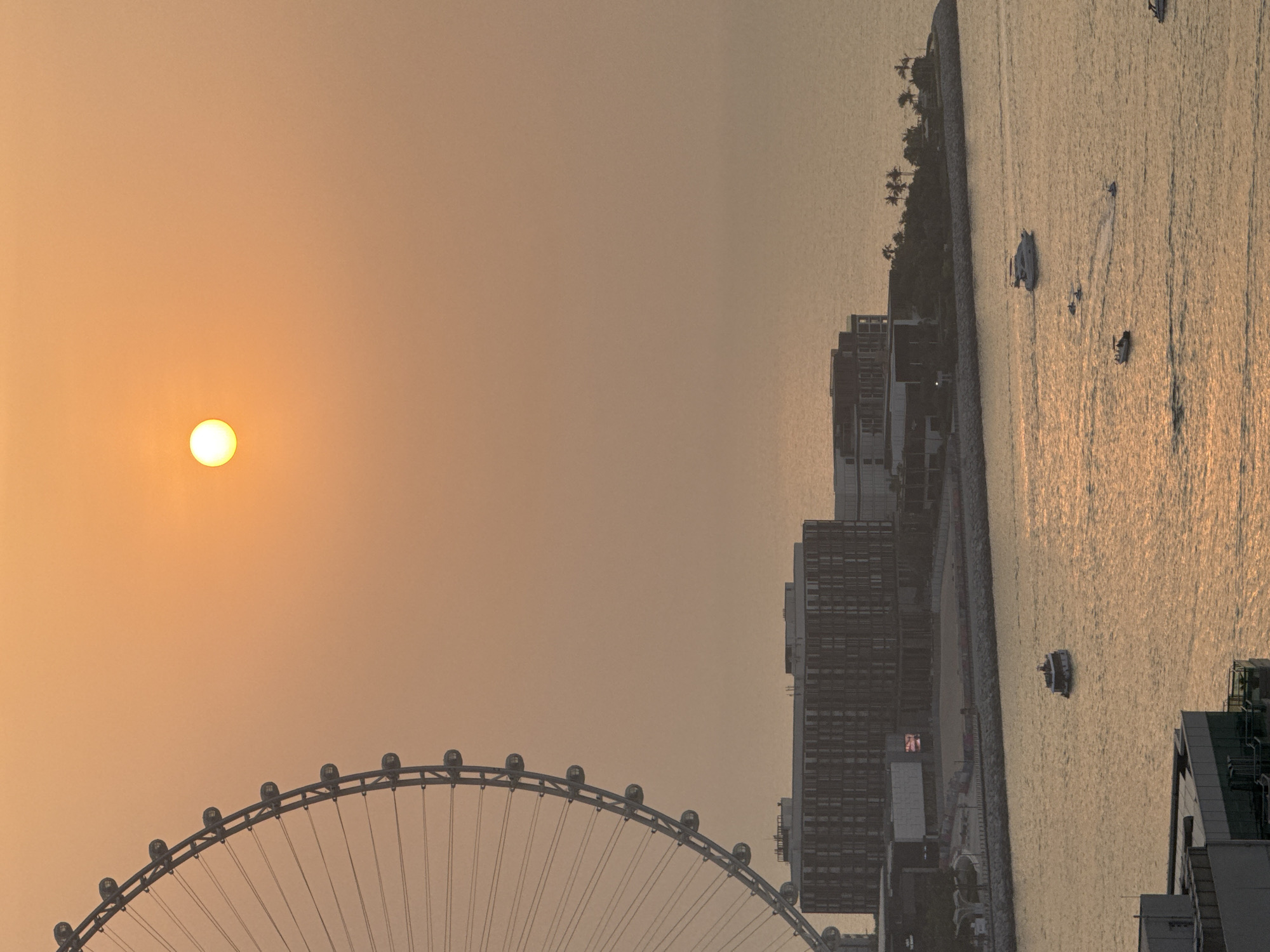 The image depicts a coastal city scene in Dubai, United Arab Emirates, during a hazy sunset. A large Ferris wheel, partially visible on the left, dominates the upper left portion of the frame. Below the Ferris wheel, several modern buildings line the waterfront, some with visible windows and architectural details. The sea occupies the middle and lower portions of the image, reflecting the warm, orange hues of the setting sun. Several boats of various sizes are scattered across the water, some moving and leaving wakes. The overall atmosphere is calm and serene, with the hazy sunlight casting a soft glow over the entire scene. The location is likely JBR, with the Ain Dubai Ferris wheel and waterfront developments clearly identifiable.