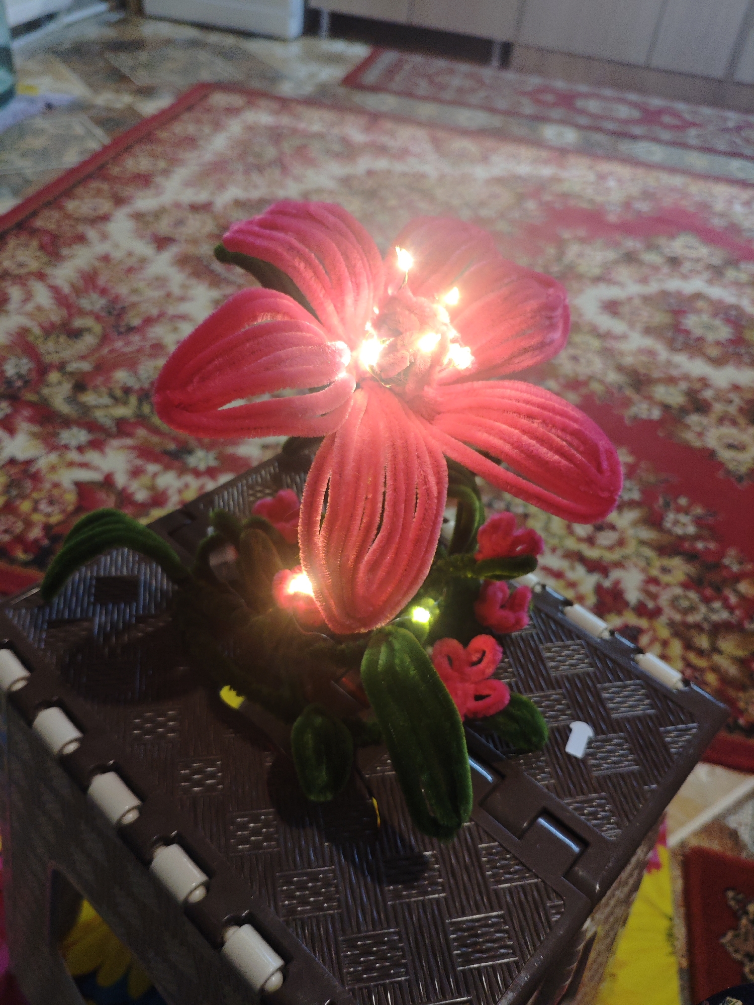 An illuminated, handmade, pink pipe cleaner flower sits on a brown plastic woven surface. The flower has multiple large petals and smaller buds, with tiny lights embedded within the petals and stem, creating a warm glow. Green pipe cleaner leaves and stems support the flower. The background consists of a patterned rug with red and beige tones, and a light-colored wall in the distance. This scene is indoors, and no specific location in Armavir, Russia can be determined from the image.
