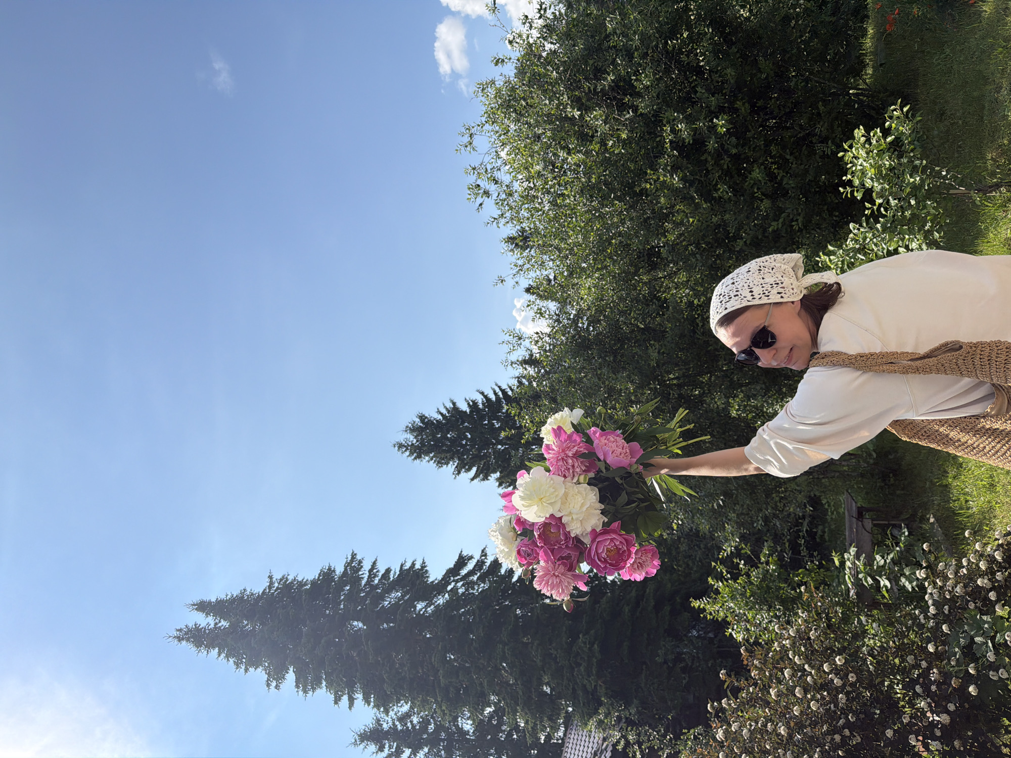 In an outdoor scene bathed in sunlight, a woman stands in what appears to be a garden or park area. She is wearing a white top and a white headscarf, along with sunglasses. In her right hand, she holds a large bouquet of pink and white peonies, lifting it above her head. A woven tote bag hangs from her shoulder. Behind her, the environment is lush with greenery, featuring tall evergreen trees and various bushes and plants, suggesting a natural or semi-rural setting. The sky above is a clear blue with a few wispy clouds. The overall impression is one of a pleasant, sunny day, possibly in a residential area or a botanical garden, given the presence of flowers and manicured landscaping. There is no direct evidence to pinpoint the specific location as Priksky, Russia, but the visual cues are consistent with a temperate climate and rural or suburban garden aesthetics.